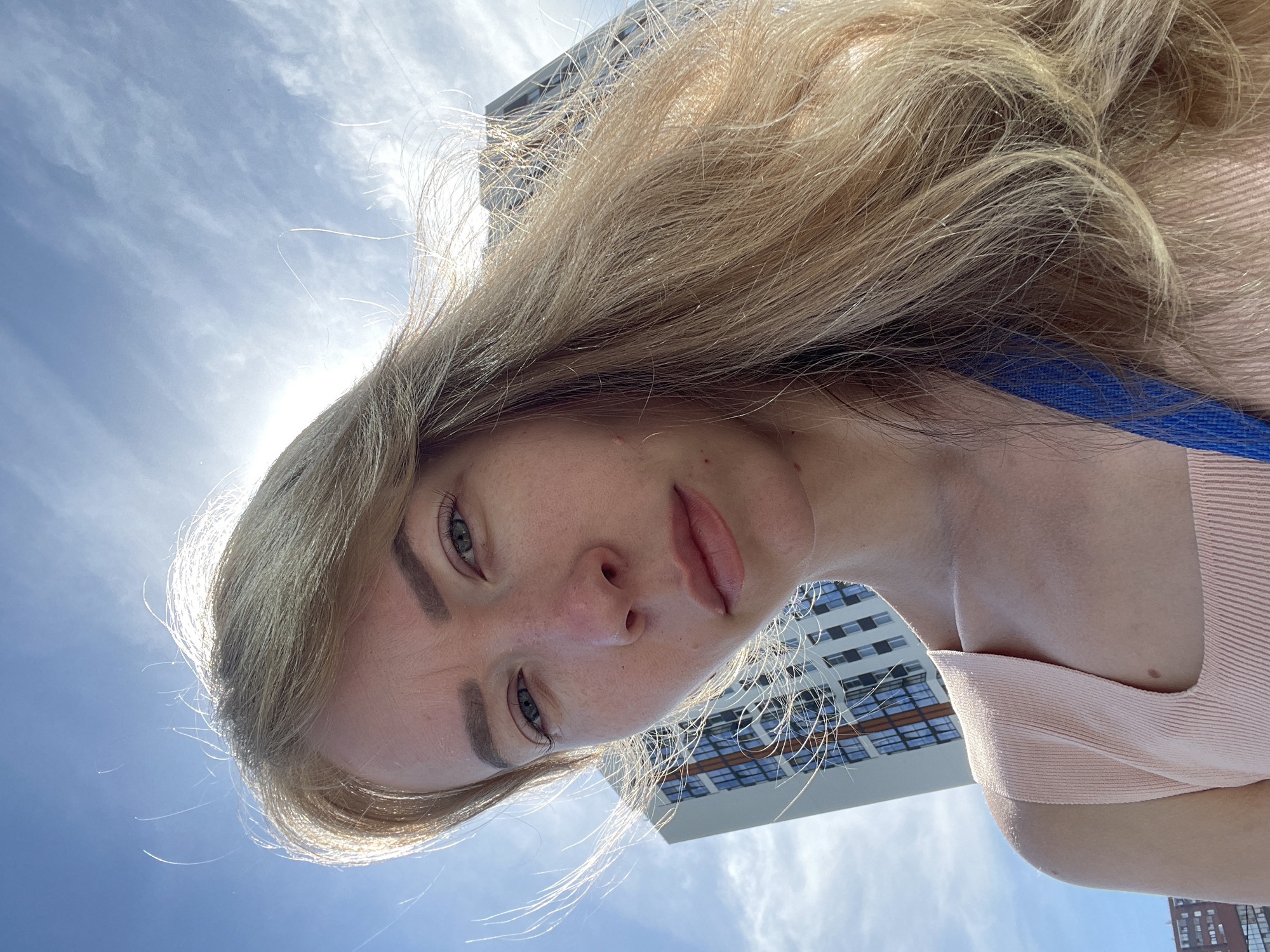 A low-angle, close-up selfie of a fair-skinned woman with light blonde hair is oriented so that the sky and a partial view of a tall building are visible in the background. The woman's face is centered, displaying light green eyes, fair skin, and freckles across her nose. She is wearing a light pink top with a blue strap visible on her right shoulder. The building features a grid-like pattern of windows and white facade. The sky is blue with scattered white clouds and the sun shining intensely, causing lens flare. This image appears to have been taken outdoors, likely in a city area based on the presence of the building. No specific location (Perm, Russia) can be determined from the image without additional information. There are no apparent actions or interactions taking place in the image.