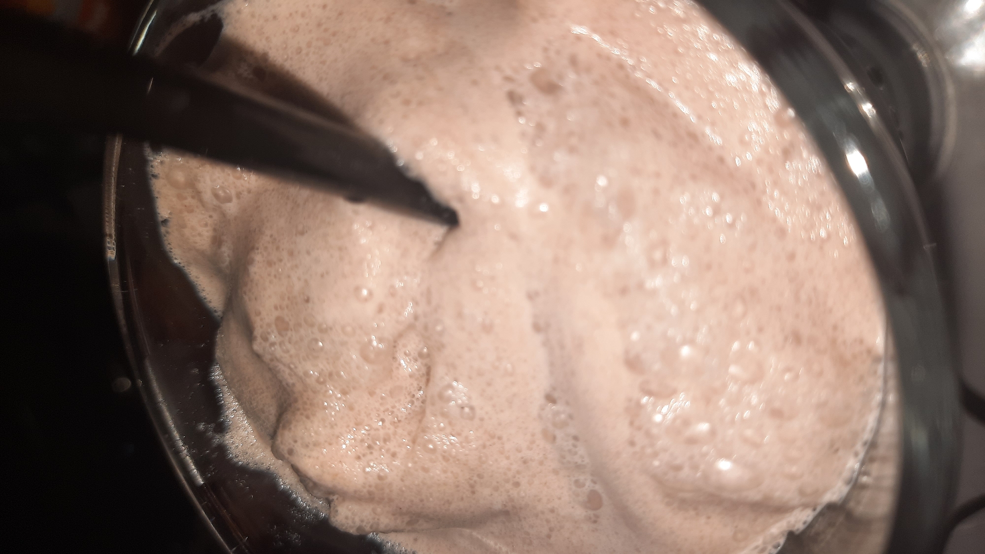 A close-up, slightly out-of-focus shot captures the frothy surface of a chocolate beverage in a clear glass. A dark straw is submerged in the drink, its tip creating ripples and disturbing the foam. The lighting is dim, with highlights reflecting off the bubbles and the rim of the glass. The background is dark and indistinct. This scene is likely taking place indoors, possibly in a home or cafe, and there are no discernible elements to place it specifically in Шклоў, Belarus.
