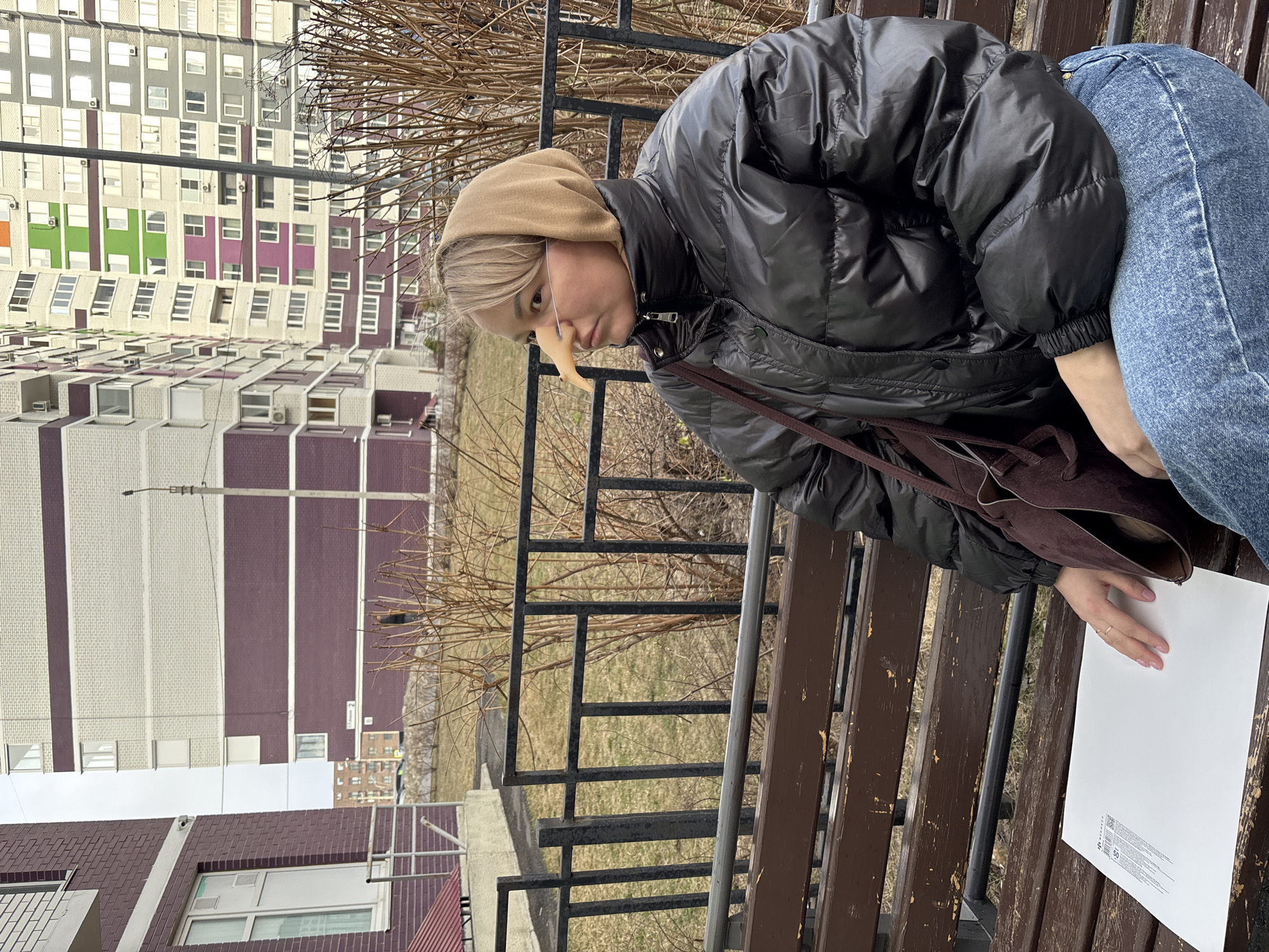 A sideways full shot depicts a person sitting on a bench outdoors. The individual wears a black puffer jacket, blue jeans, and a light brown head covering. They have what appears to be a false, elongated nose attached to their face. A brown bag hangs from their shoulder, and they are holding a white piece of paper on the bench. The background shows a grassy area behind a black metal fence, and multiple-story apartment buildings in various colors are visible in the distance. The location is identified as Izhevsk, Russia.