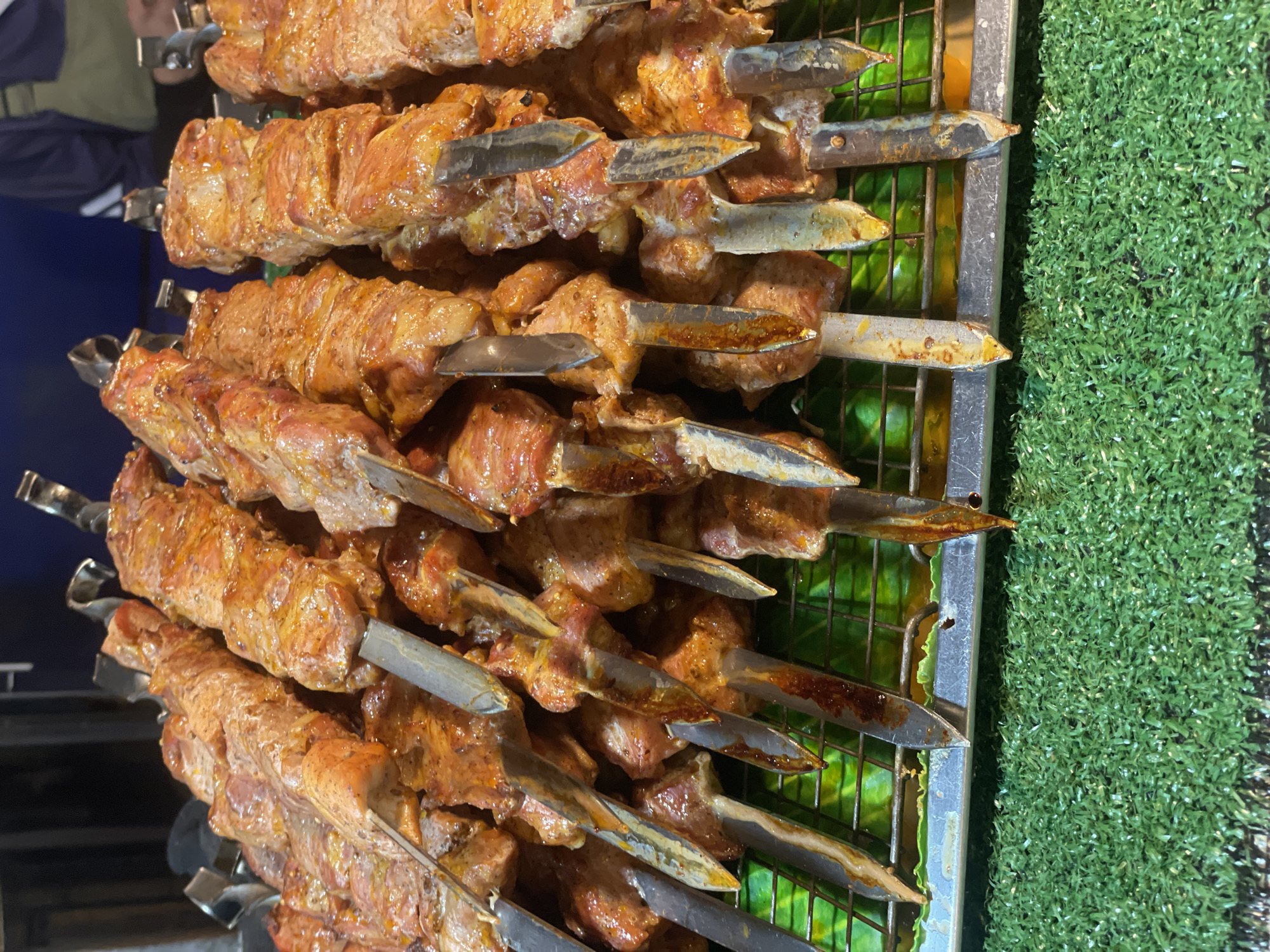 A close-up, eye-level, outdoor shot shows a rack of beef skewers on metal skewers, arranged diagonally from the bottom left to the top right. The skewers are resting on a green, grate-like surface, which is itself on a bed of artificial green grass. The beef is reddish-brown with visible fat marbling and appears to be seasoned or marinated. Some skewers are more densely packed with meat than others. The background is dark and blurred, but hints of other food stalls and possibly people can be discerned. The scene suggests a street food vendor or market setting, likely in Pattaya City, Thailand, given the bright, humid atmosphere often found there, where such grilled meats are a popular culinary offering.