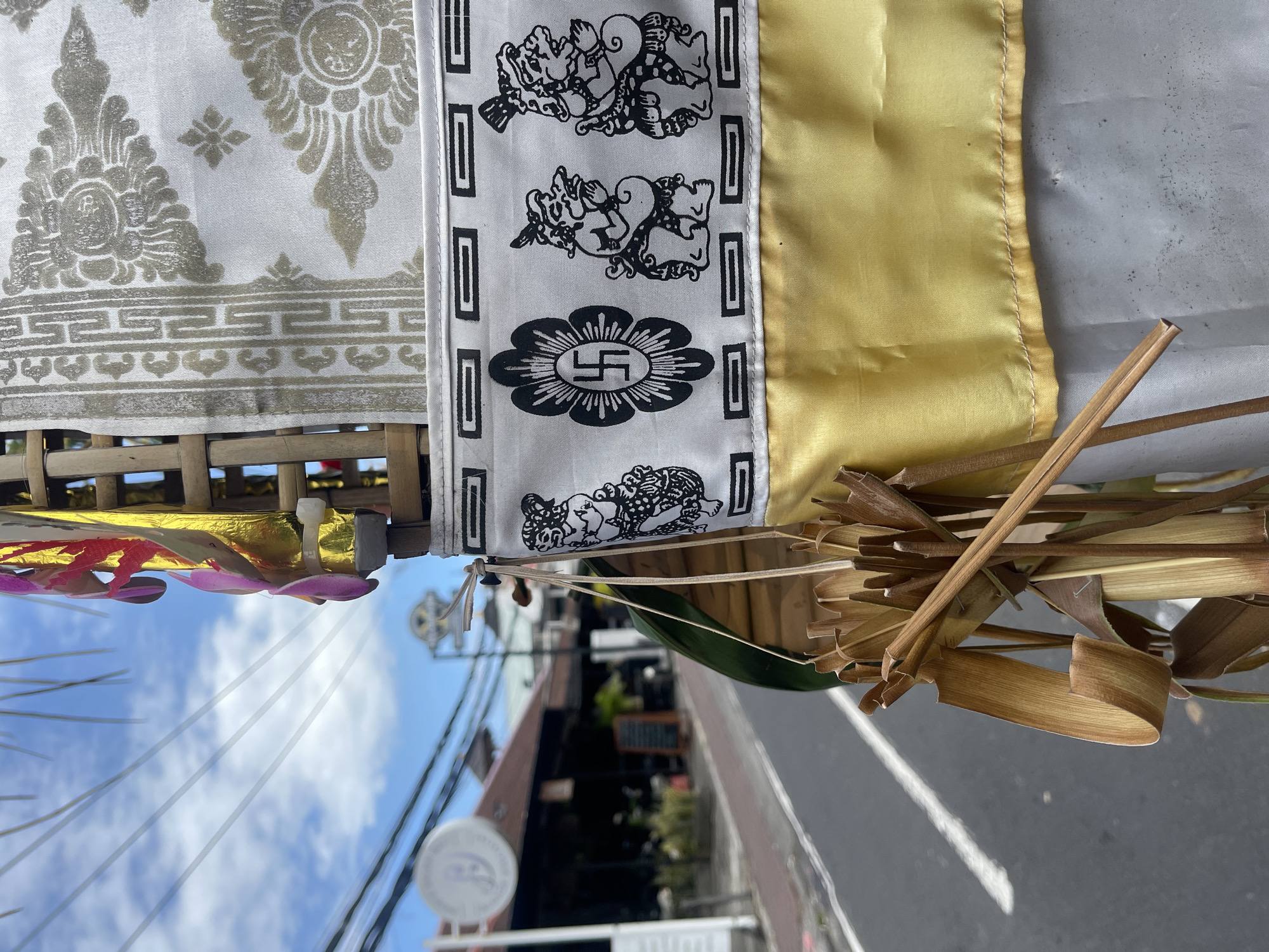 In Kuta, Indonesia, a close-up shot captures a section of a decorative bamboo structure adorned with white fabric featuring black, intricate patterns. The fabric displays a border of repeated rectangular shapes, followed by a central motif of a swastika within a floral design, and then a sequence of three stylized human figures in prayer. Below this, a vibrant yellow sash is attached. In the foreground, dried palm fronds and bamboo elements are visible, hanging from the structure. The background reveals a street scene with a paved road and sidewalk, lined with shops and buildings. The sky above is a bright blue with scattered white clouds. Power lines crisscross the frame against the sky.
