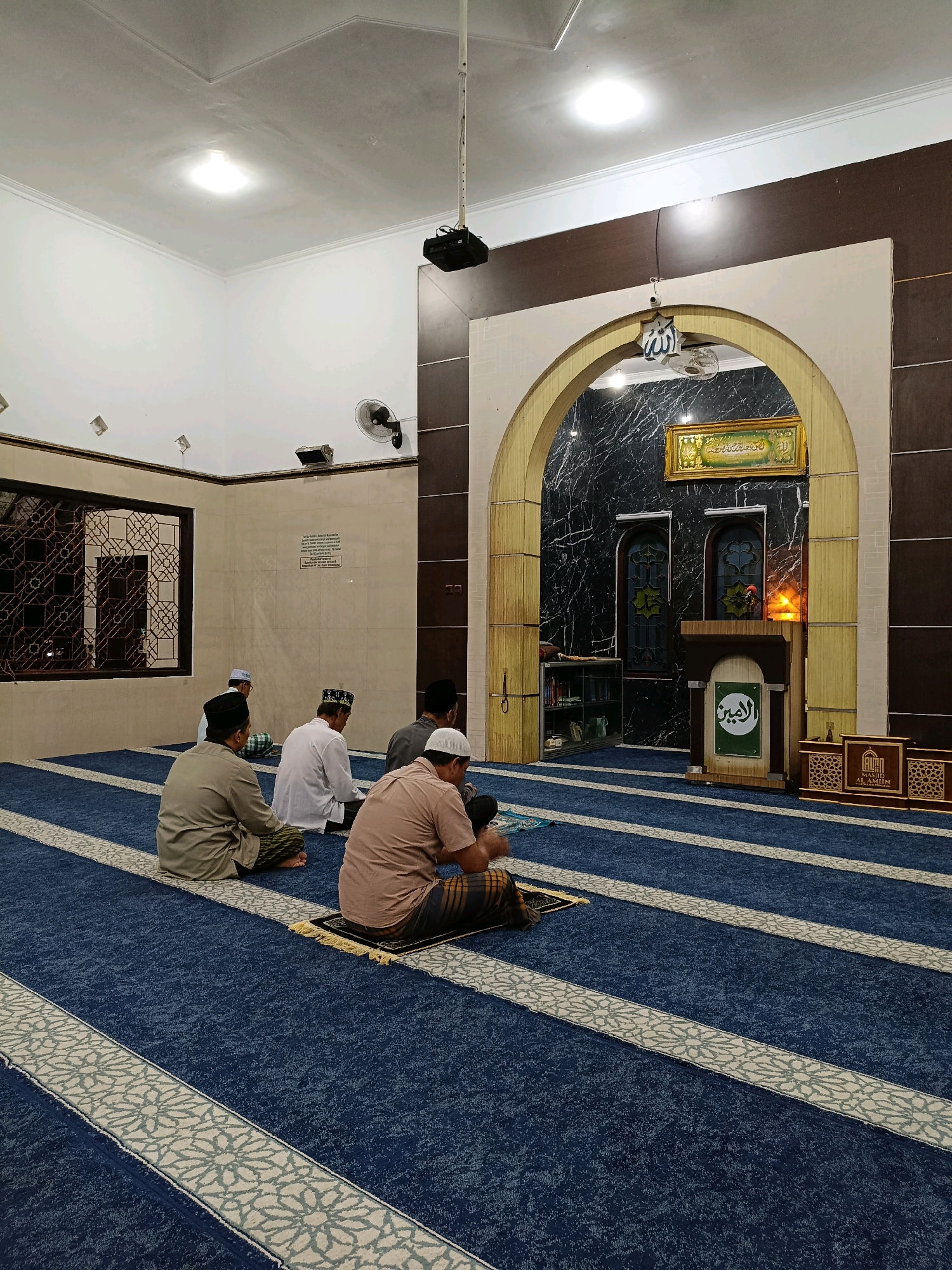 The image depicts the interior of a mosque, likely located in Maguwoharjo, Indonesia, with several men seated on a patterned blue carpet, facing the mihrab. The room is brightly lit by overhead lights and has white walls with a beige trim. A large arched opening leads to the mihrab area, which is decorated with dark marble-patterned walls and features a podium with Islamic calligraphy and a decorative screen. There's a framed artwork above the arch, likely a Quranic verse. To the left, a large window with intricate geometric patterns is visible. The men are dressed in traditional attire, with some wearing peci caps. They appear to be engaged in prayer or a religious gathering. The overall atmosphere suggests a peaceful and solemn occasion.