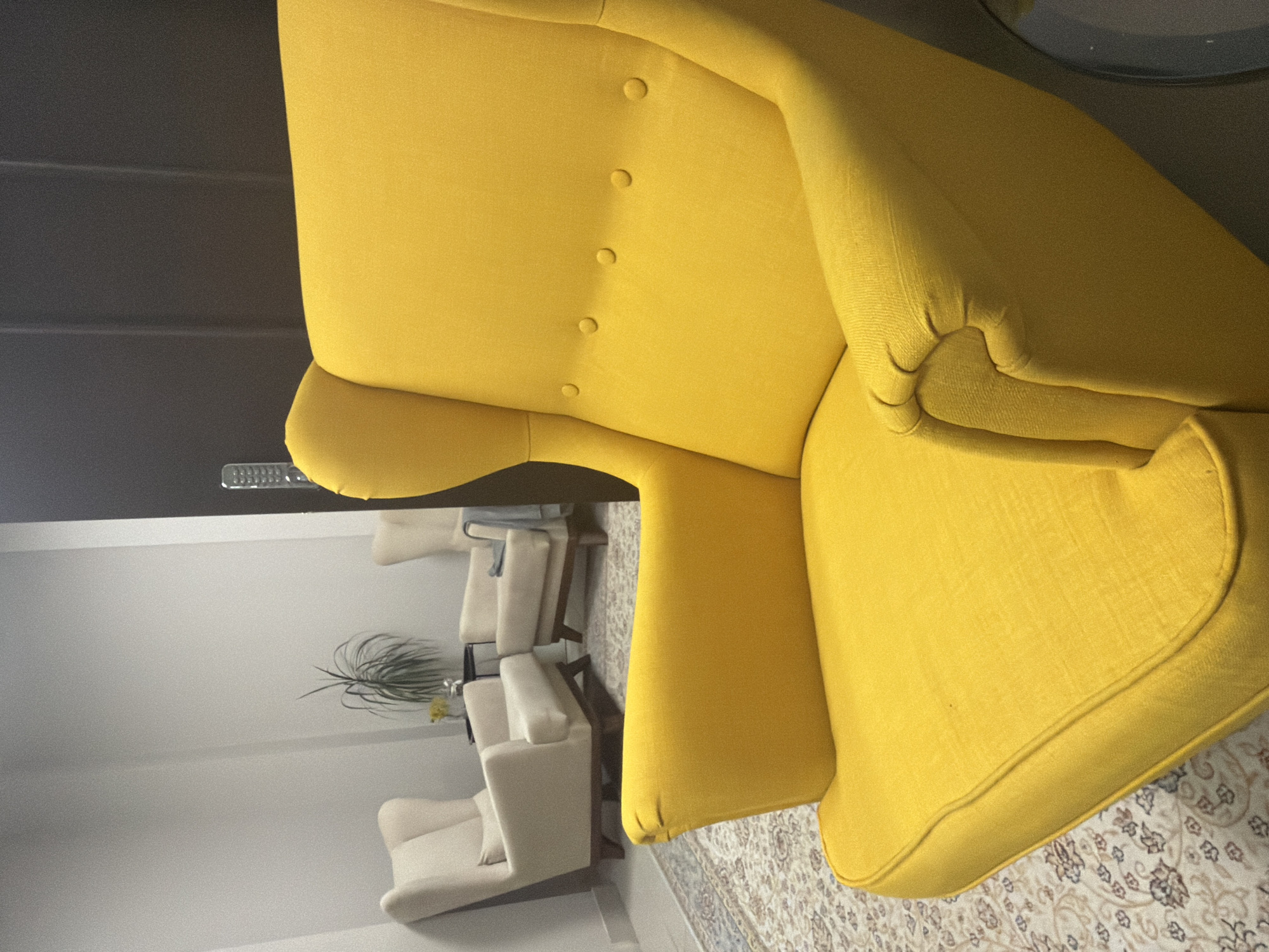 The image shows the corner of a room with a yellow armchair in the foreground. The armchair has buttons on the upper back and a rounded seat. The floor is covered with a patterned rug. In the background, there are several white armchairs and a plant on a white surface. The location is likely Perm, Russia, based on the presence of furniture and interior design elements.