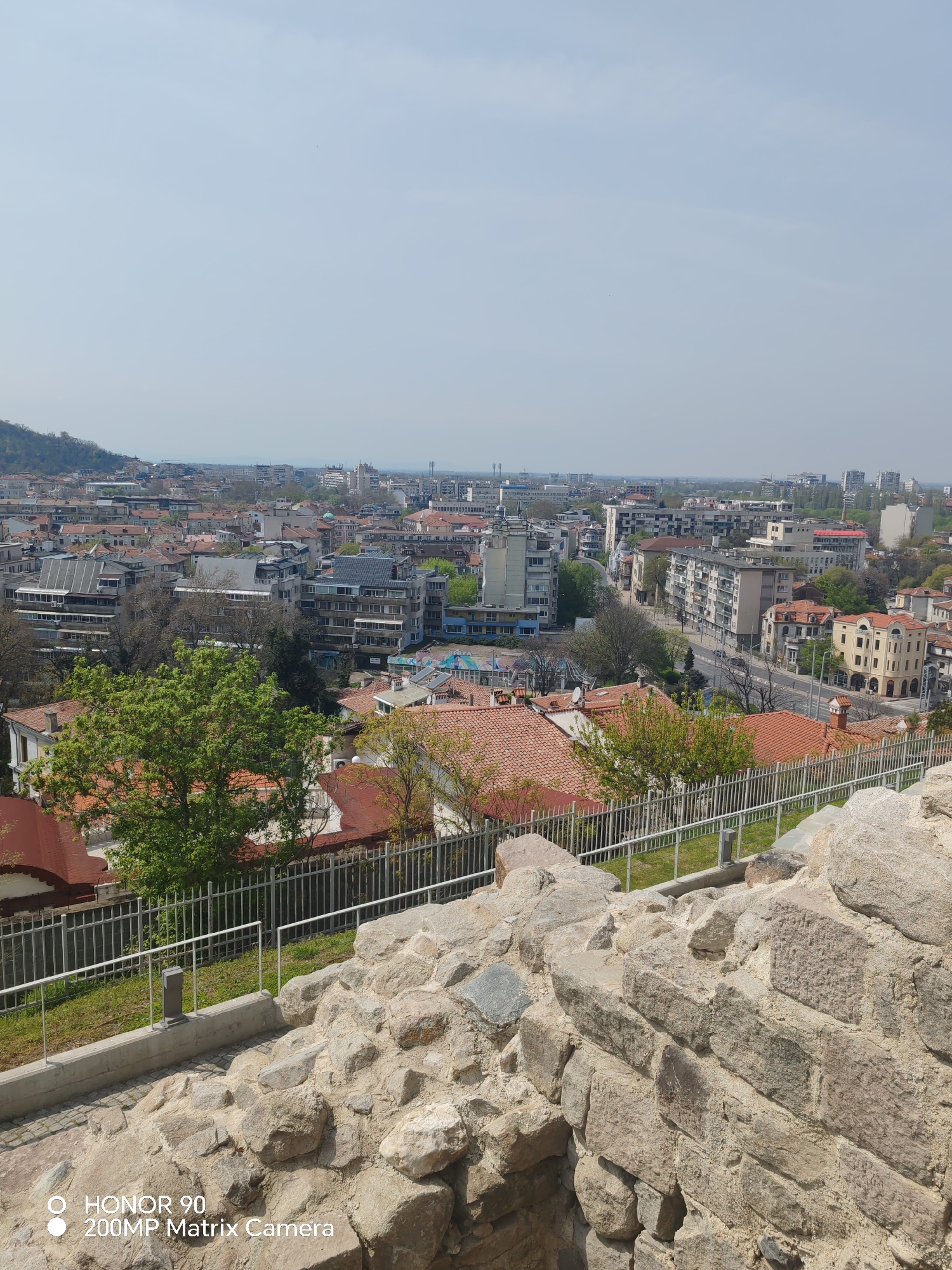 The image shows a panoramic view of Plovdiv, Bulgaria, captured from Nebet Tepe, one of the city's ancient hills. The foreground features a stone wall, part of the archaeological remains of the Thracian settlement, offering a clear overlook. A modern metal railing runs along the wall's edge, delineating the observation area.
The city unfolds beneath, with a mix of residential and commercial buildings visible, primarily constructed in various architectural styles from the mid-20th century to modern times. Red tile roofs are prominent, contrasting with the concrete and glass facades of the taller structures.
In the midground, streets and avenues are seen, with sparse vehicle traffic visible. Mature trees provide a green canopy interspersed with the buildings. The skyline of Plovdiv is moderately dense, with buildings of varying heights, and is set against a hazy sky. A mountain appears to the left of the frame. The picture was taken during the day, with diffused light.