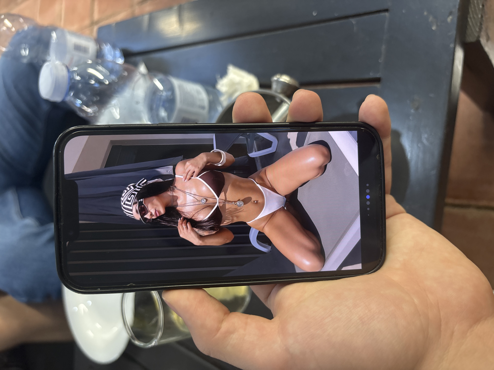 A right hand holds a black smartphone, displaying an image of a woman. The woman is kneeling on a grey lounge chair or sunbed, posed with her body slightly angled. She wears a dark brown and white two-piece bikini, a black and white geometric pattern headscarf, and dark sunglasses. Multiple silver-toned necklaces with pendants, a belly button piercing, and a white bangle bracelet are visible accessories. Her skin appears bronzed, with highlights on her knees and shoulders. Behind her in the phone's image are dark, vertically slatted panels. The phone screen also displays navigation dots at the bottom, with one illuminated blue, indicating the current image in a sequence.
Outside the phone, the scene is set on a dark, possibly slatted, outdoor table. A clear glass containing a yellowish liquid is positioned to the left of the phone. Two out-of-focus plastic water bottles with white caps and a white ceramic dish are visible in the background on the table. A person wearing blue denim jeans is seated in the blurry upper background. The ground beneath the table is composed of terracotta-colored tiles. The overall environment suggests a casual outdoor setting during daylight hours, in the city of Blagoevgrad, Bulgaria.