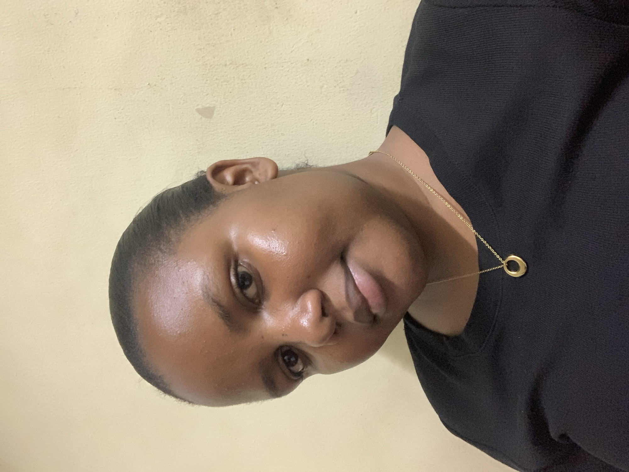 A close-up, angled headshot of a Black woman against a pale yellow wall. She is wearing a black top and a gold necklace with a circular pendant. No evidence suggests the location is Dar es-Salaam, Tanzania.