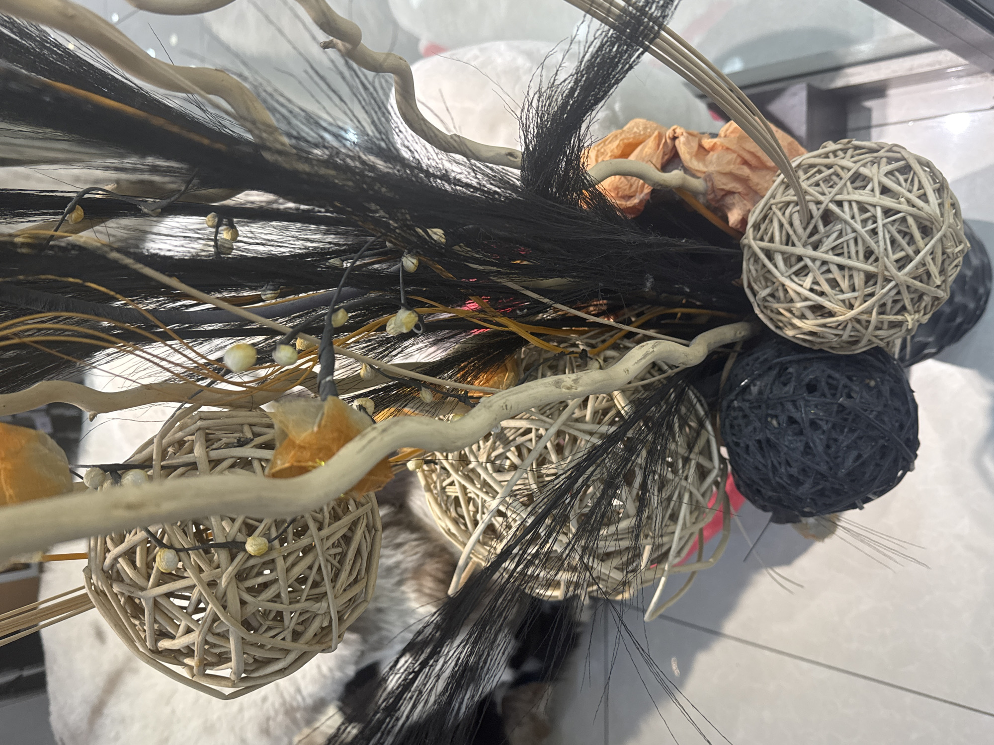 The image displays a close-up, eye-level view of a decorative arrangement. The arrangement consists of several spherical objects made of woven natural materials, appearing as balls of rattan or similar fibrous plants. These balls are of varying sizes and colors, including a dark grey one, a light beige one, and another lighter, larger one with a more open weave. Interspersed among the balls are dried, twig-like branches, some of which are smooth and light-colored, while others are darker and more irregular. There are also wispy, dark strands, possibly feathers or fine plant material, adding texture and depth. Small, bead-like accents in a pale yellow color are visible, strung on thin wires. Some dried, crinkled elements, appearing floral or leaf-like and in a muted orange hue, are also present within the arrangement. The background is blurred but suggests an indoor environment with soft, possibly furry, textured surfaces and a tiled floor. There is no indication of the city of Bishkek City, Kyrgyzstan, or any specific location. The image is static and focuses solely on the decorative object.