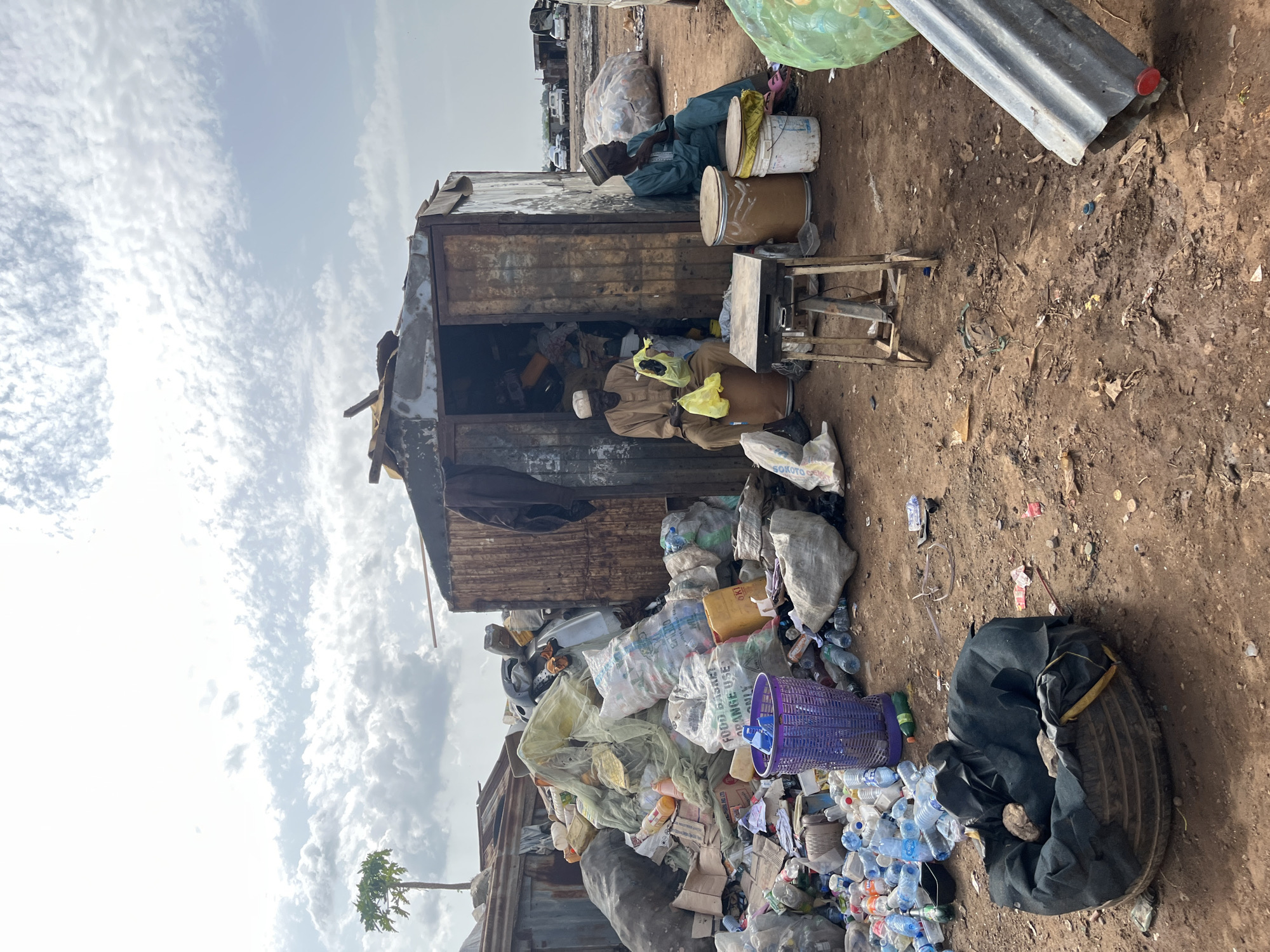 The image depicts a scene of a recycling or waste collection area in Jos, Nigeria. In the foreground, a large pile of sorted and unsorted waste, including plastic bottles, bags, and other debris, is visible. Two men are seated outside a makeshift shelter constructed from corrugated metal. One man, wearing a light brown garment and a cap, is sitting on a barrel inside the shelter's entrance, holding a yellow plastic bag. The other man, dressed in a blue garment and a patterned cap, is seated on a white bucket to the right of the shelter, looking away from the camera. In front of the shelter, there's a small metal stand with various items on it. The environment appears to be a dusty, unpaved area with more piles of waste and some vehicles visible in the background. The sky is cloudy.