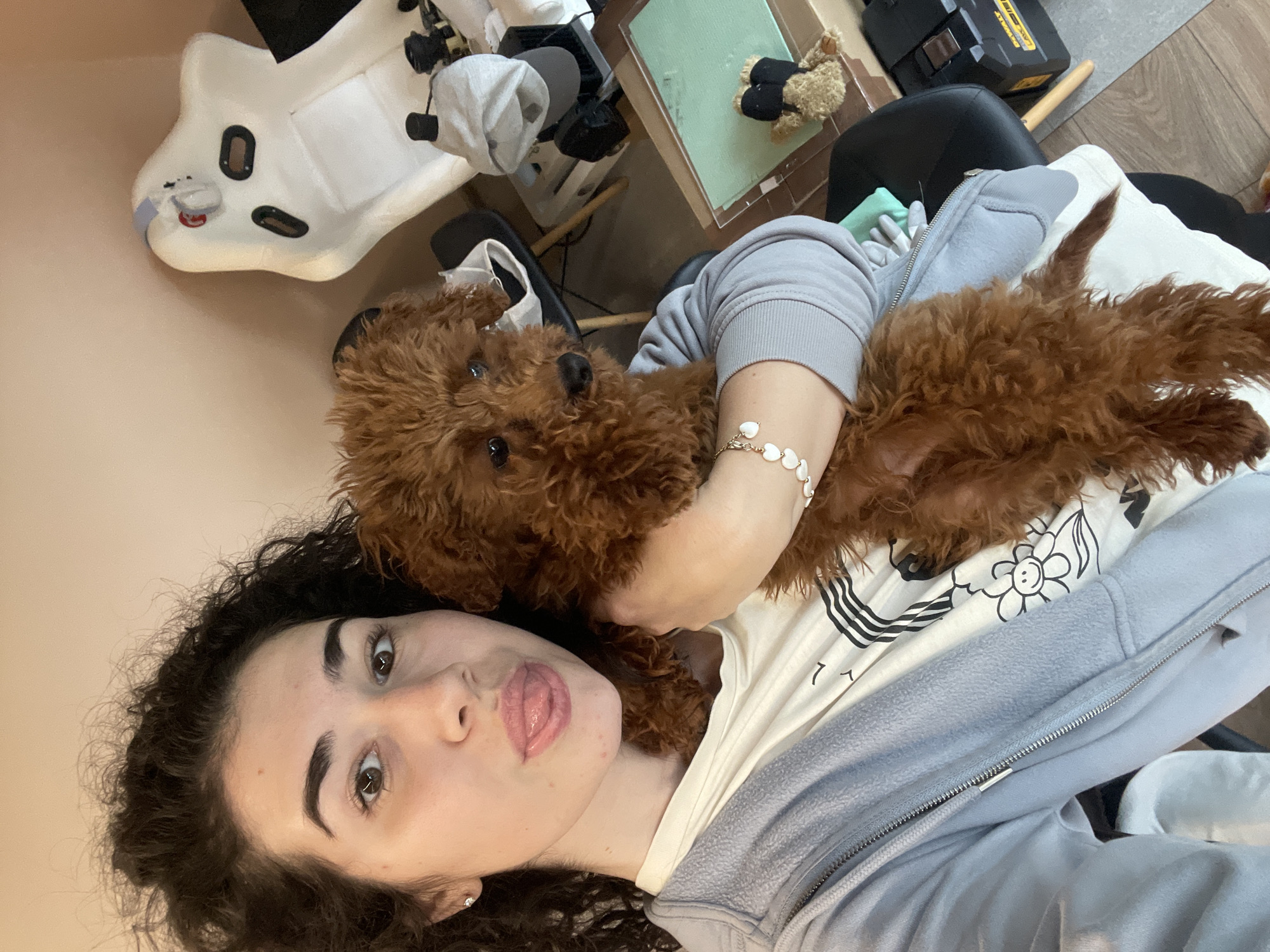 A woman with dark curly hair lies on her back, cradling a small, brown poodle. The woman has fair skin, dark eyebrows, and is sticking out her tongue. She wears a white t-shirt with a black flower design and a light grey zip-up jacket. A bracelet of white heart-shaped beads is visible on her arm. The dog is curled up against her, with its face close to hers. The background includes a white baby bouncer seat, a table with a small teddy bear, a black office chair, and a wooden floor. It is not possible to determine the location in the city of Sofia, Bulgaria from the visual information.