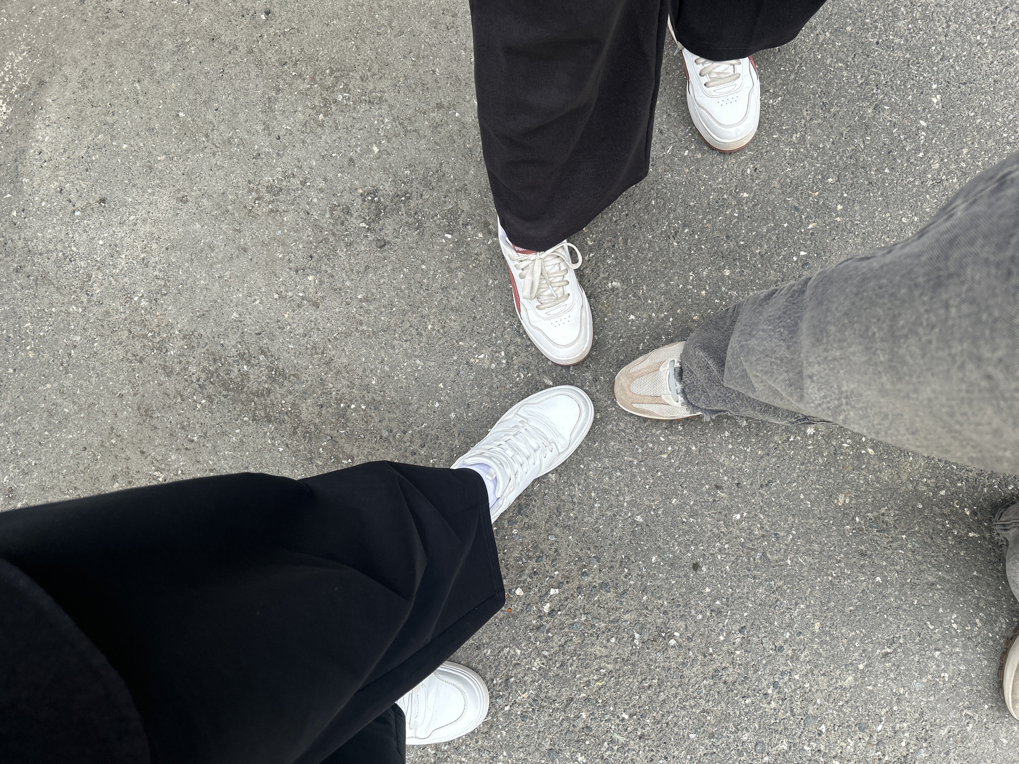 The image shows a close-up, overhead view of three pairs of feet standing on a grey, pebbled asphalt surface. The feet are clad in various sneakers and pants. On the left, a person wearing black pants and white sneakers is visible. In the upper right quadrant, another person is wearing black pants and white sneakers with red accents. The third person, in the lower half of the image, is wearing grey jeans and light brown/tan sneakers. The scene appears to be outdoors, likely a sidewalk or street, with no specific landmarks to indicate a precise location within the city of Perm, Russia. The lighting suggests it could be daytime.