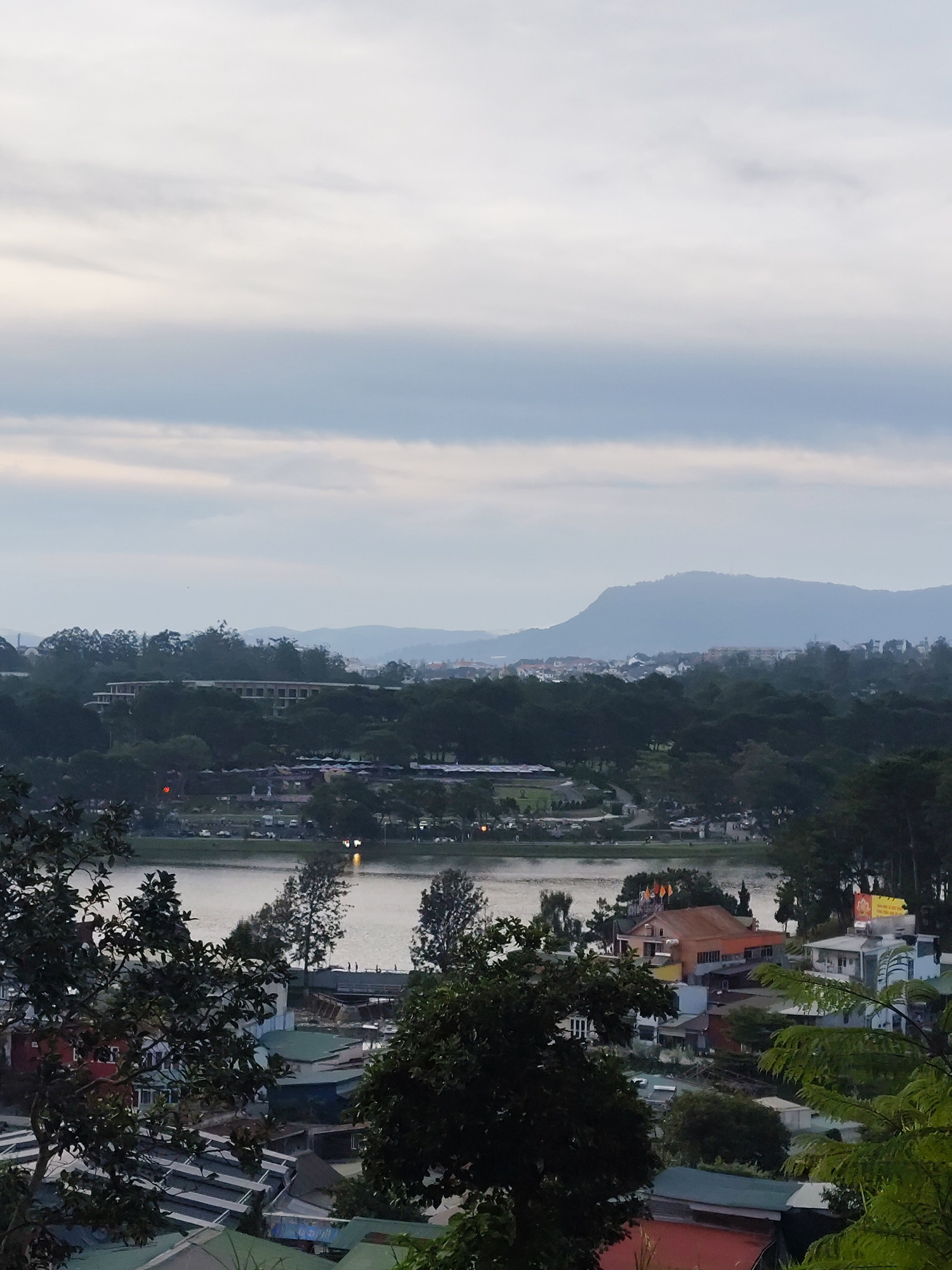 This is an elevated, wide-angle, vertical shot of Xuan Huong Lake in Da Lat, Vietnam, during overcast weather. In the foreground, dark green foliage obscures the lower portion of the frame. The middle ground features the tranquil, light-brown water of Xuan Huong Lake. Along the lake's edge, a paved walkway is visible, with trees and scattered parked vehicles. Beyond the walkway, there is a dense expanse of dark green trees. Further back, a large, elongated building with a light-colored facade and numerous windows is situated amidst the trees. In the distance, rolling hills and mountains, softened by the atmospheric haze, form the backdrop. The sky is overcast with large, horizontal bands of gray and white clouds. The scene depicts a serene, natural landscape integrated with urban elements.