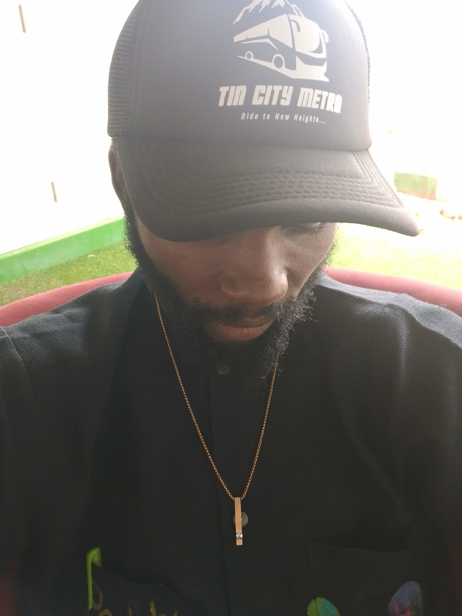 In a close-up, medium-shot portrait, a person wearing a black trucker hat with "TIN CITY METRO Ride to New Heights..." embroidered in white, and a simple gold chain with a rectangular pendant, looks downwards. The person has a dark complexion, a short beard, and is wearing a black collared shirt. The background is out of focus, showing a blurred green and brown area, possibly indicating an outdoor setting with vegetation and a red chair. The lighting suggests it is daytime, with a bright light source to the left of the frame, creating some highlights and shadows. The overall impression is a casual, personal snapshot.