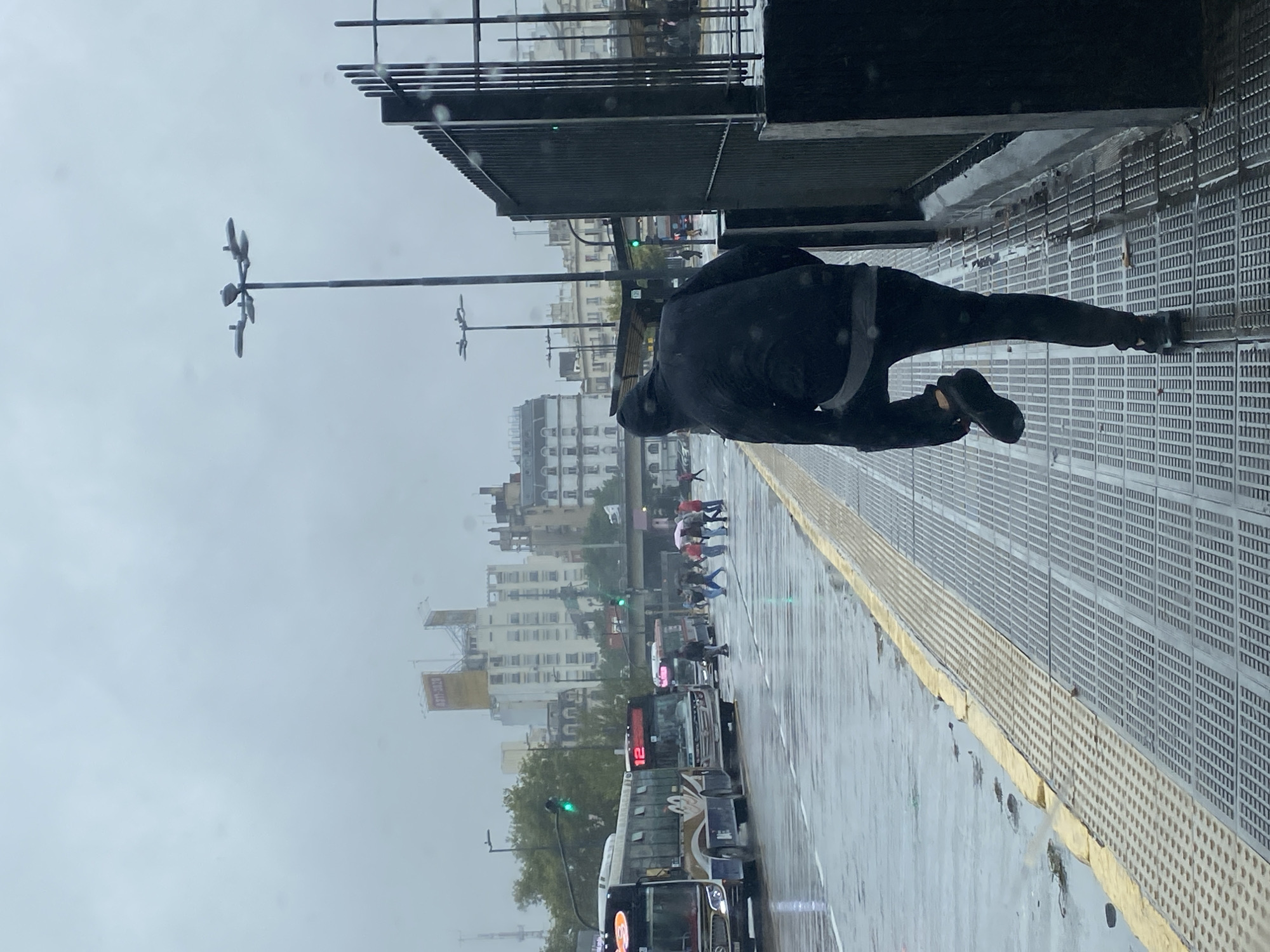 The image captures an urban street scene on an overcast, rainy day, viewed from an elevated perspective, possibly a pedestrian bridge or overpass. In the immediate foreground, a person dressed in dark clothing, including a jacket and pants, is seen from behind, walking on a textured, grey grated metal platform. This platform features a distinctive yellow stripe along its edge.
Below the elevated platform, a street is visible, appearing wet and reflective from recent rain. In the middle distance, several pedestrians are crossing the street, some holding colorful umbrellas, indicating ongoing rain. A public bus, prominently displaying the route number "12" on its front and side, is present on the wet road. Other vehicles are also seen, and green traffic lights are visible, indicating active traffic flow.
The background reveals a dense cityscape composed of numerous multi-story buildings, typical of an urban setting. The sky is uniformly grey and overcast, contributing to the somber weather atmosphere. Visible raindrops or streaks, possibly on the camera lens or a window, further emphasize the rainy conditions. In the far background, a yellow banner or sign on a building or construction site displays the text "1978 - 2028".
The presence of the "12" bus route is a strong location cue, aligning with public transport systems found in Buenos Aires, Argentina, as per the provided context. The scene depicts everyday life in a bustling city during inclement weather, without conveying specific emotions beyond the general mood set by the weather.