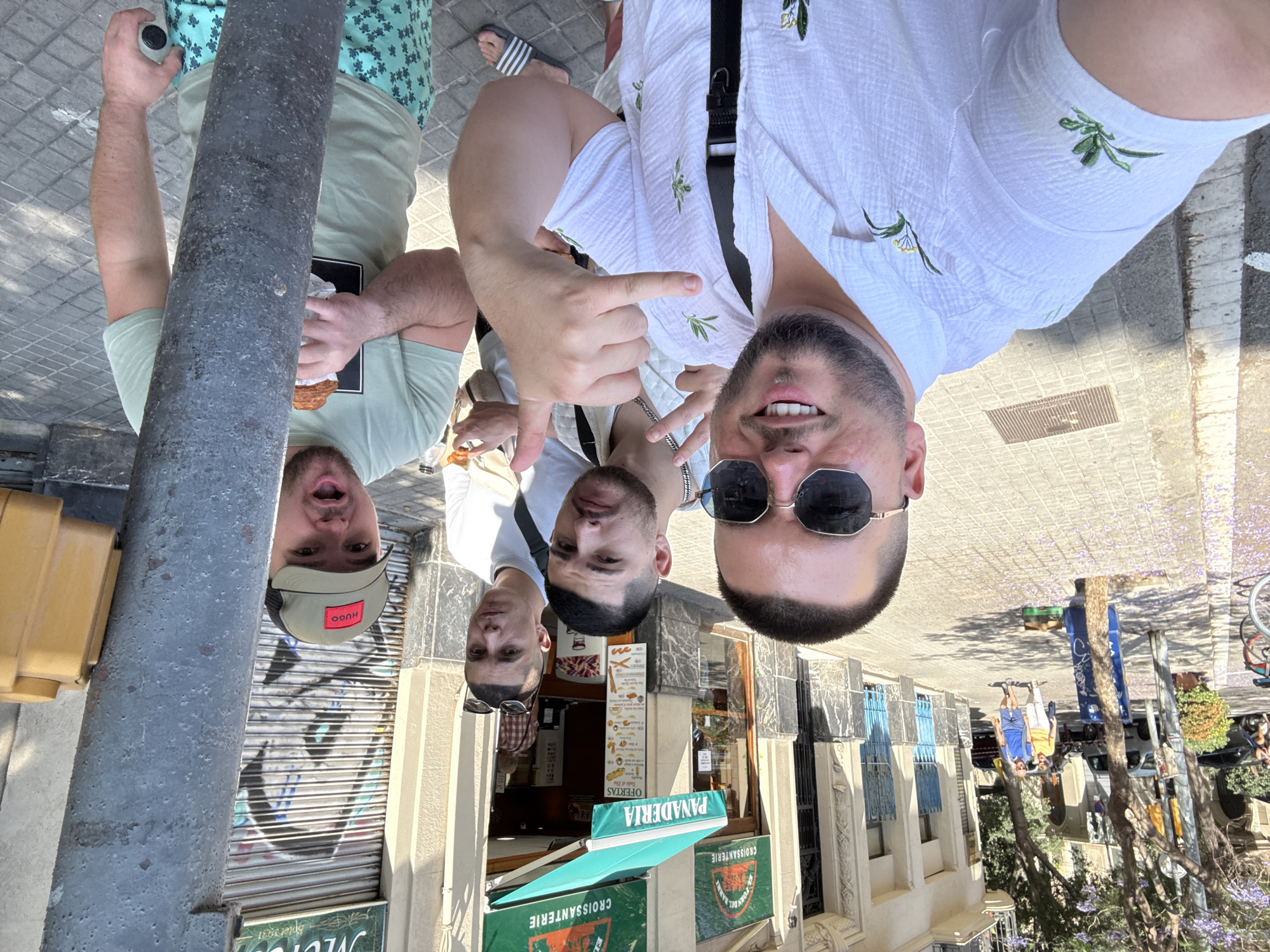 Four men are in a selfie photograph. The man in the foreground is wearing a white shirt and sunglasses, and he is making a hand gesture. Behind him and to his left are two more men, one in a white shirt and the other with sunglasses. The man to the far left is wearing a green shirt, a cap, and eating something. A street lamp is positioned to the left of the men. Behind the men is a building with a storefront labeled "Panaderia Croissanterie." The awning of the shop is green and white. In the background is a street with trees and buildings, indicating a city setting. Based on the language on the storefront, the location is likely Barcelona, Spain.