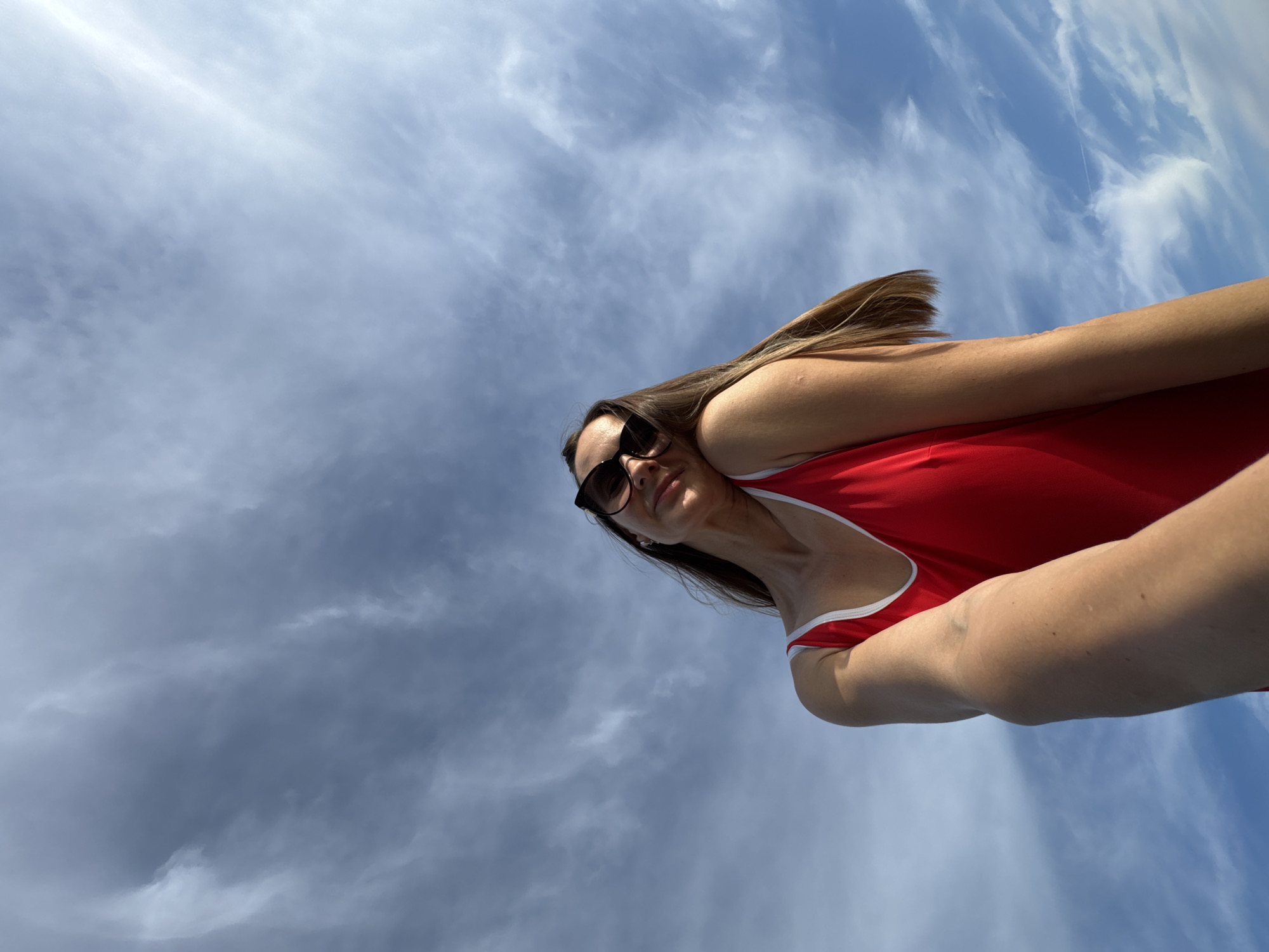 A woman in a red swimsuit and sunglasses is looking up at the sky. The sky is blue with white clouds. The image appears to be taken outdoors in Sochi, Russia.