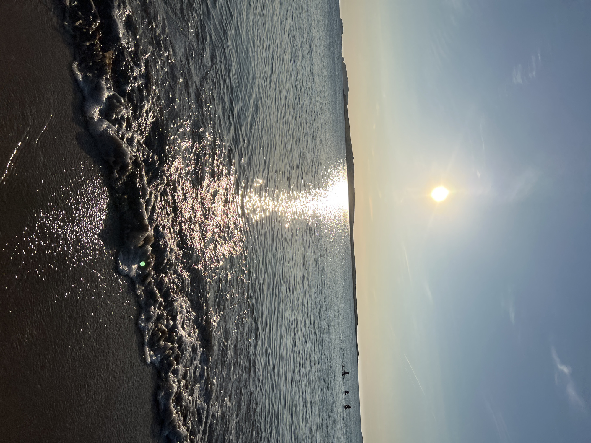 This is a low-angle, eye-level shot taken on a sandy beach at Lunniaghbeg, Ireland. The sun, positioned high in the sky and slightly to the left, casts a brilliant reflection on the water, creating a shimmering pathway. The ocean stretches out to the horizon, with a distant landmass visible on the left. In the mid-ground, three figures are in the water, appearing to be swimming or wading. Gentle waves are rolling onto the shore, creating foamy white crests as they meet the wet sand. The sky is a clear blue, with a few wispy clouds and contrails from aircraft visible. The overall impression is one of a serene and sunny day at the beach.