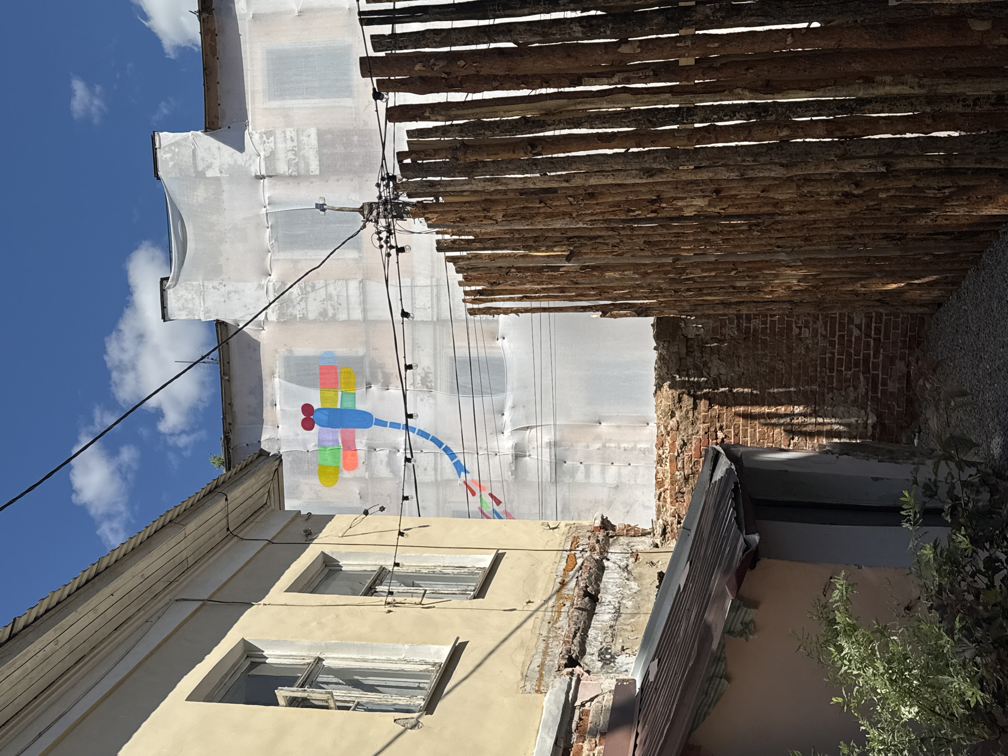 This is an outdoor, medium-shot, eye-level photograph taken in the city of Yelets, Russia. The scene depicts an alleyway between two buildings. The building on the left is old, with peeling yellow paint and exposed brickwork. It has two windows with white frames and partially open shutters. The building on the right is under renovation and covered in white sheeting, with a colorful mural of a dragonfly painted on the sheeting. A wooden fence made of vertical logs runs along the right side of the alley, partially obscuring the renovated building. Overhead, power lines crisscross the sky, and a clear blue sky with scattered clouds is visible. The ground of the alley is covered in gravel. In the foreground, a small, older structure with a corrugated metal roof is visible on the left, with some green foliage in front of it. The lighting suggests it is daytime, with sunlight casting shadows.