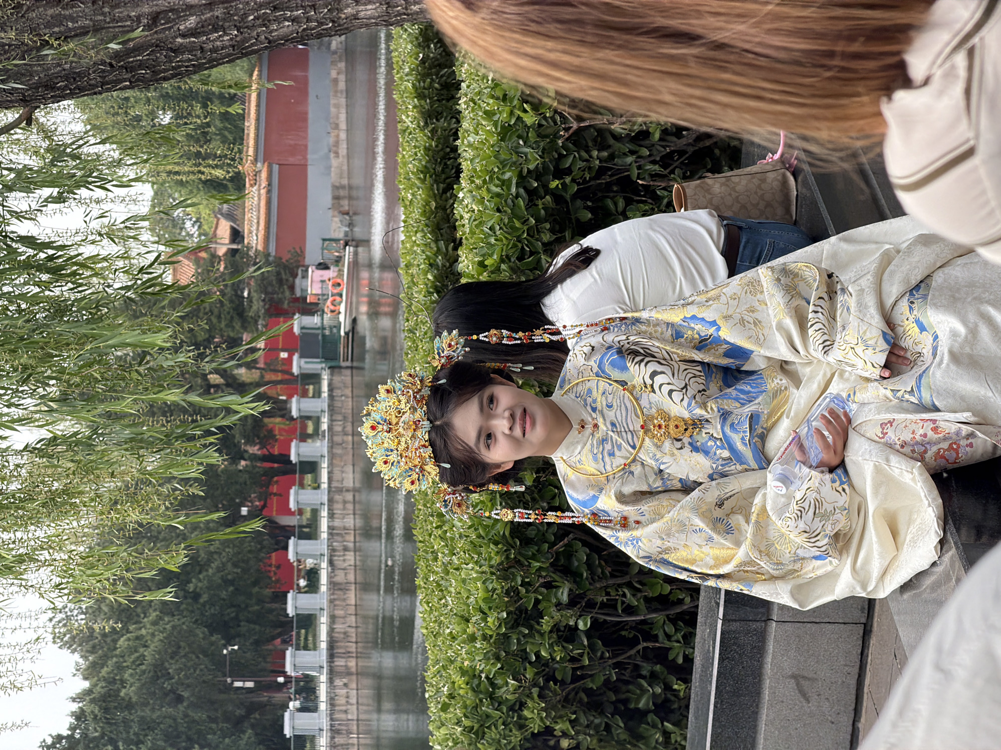 A medium shot captures a woman adorned in an ornate, traditional Chinese hanfu dress and headpiece, seated outdoors in the city of Dongcheng District, China. The attire is predominantly white, with intricate gold and blue detailing. The headpiece is golden and features elaborate embellishments. The woman gazes towards the camera with a gentle smile.
In the background, a body of water is visible, lined with a decorative stone railing and lush greenery. The water appears to be calm, reflecting the surroundings. Behind the railing, trees and a building with a red facade can be seen.
Other people are present in the scene, though less prominently featured. One individual is seated nearby, wearing jeans and a white top, with a handbag placed on the bench. A portion of another person is visible on the right side of the frame. The overall environment appears to be a park or garden-like setting, likely near a historical site or landmark within Dongcheng District.