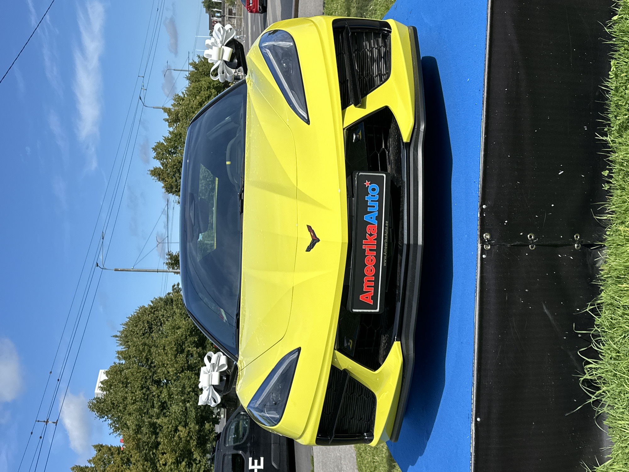A bright yellow Chevrolet Corvette is parked on a blue display platform in front of a black stage in a grassy area. The car has two large white bows attached to its side mirrors. A license plate on the front of the car reads "AmeerikaAuto" with a website address below it. The scene appears to be outdoors on a sunny day, with trees, power lines, and buildings visible in the background. The area appears to be a public space, possibly a dealership or event venue in Tallinn, Estonia. There are no people visible in the image.