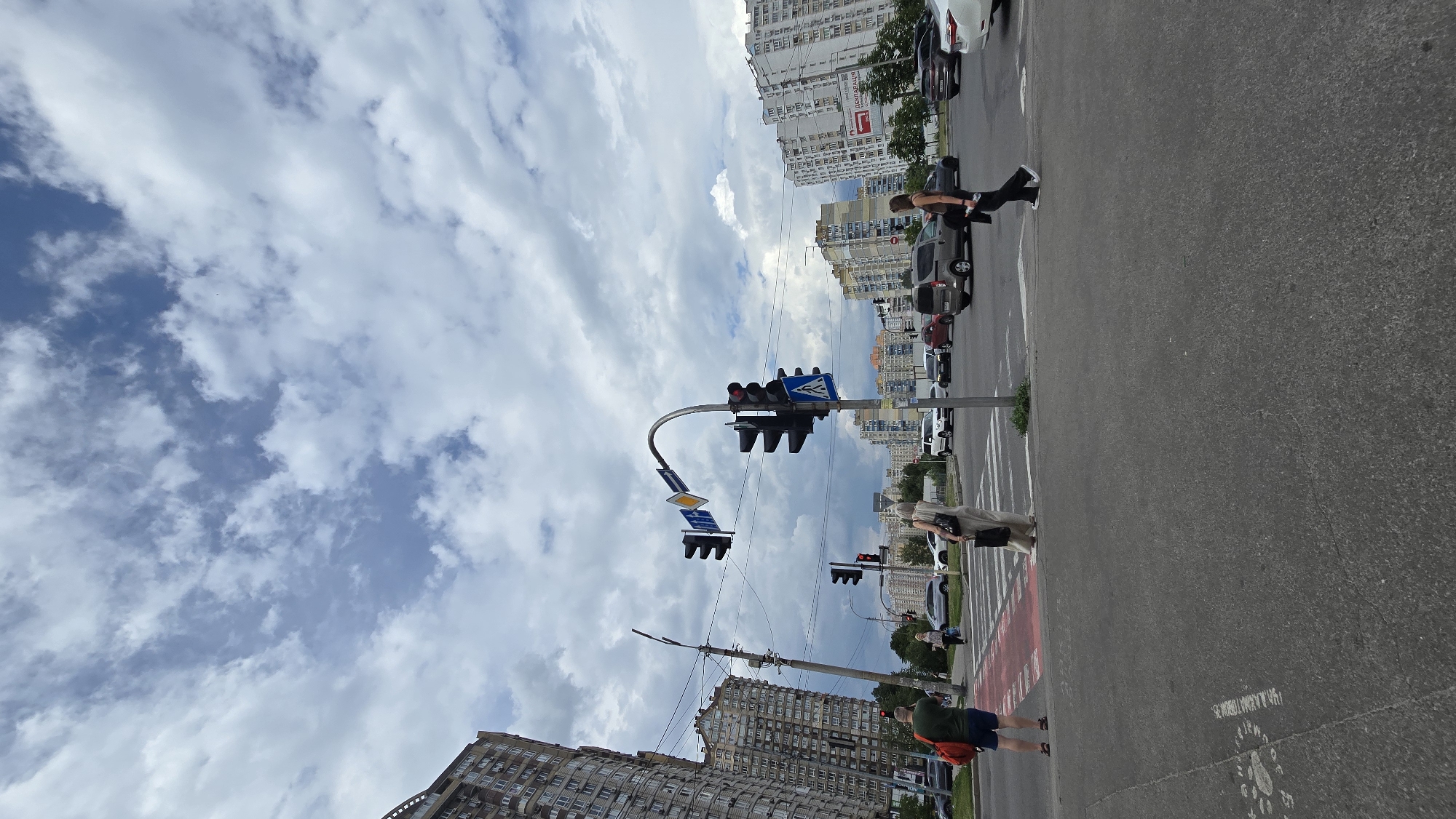 This is an urban street scene captured during daytime at the intersection of Vasylkivska Street and Holosiivskyi Avenue in Kyiv, Ukraine. The sky, occupying the upper two-thirds of the image, features a mix of scattered white cumulus clouds against a patches of blue sky.
On the left, a tall, multi-story residential building with a distinctive curved, wave-like facade dominates the background, characterized by numerous balconies and air conditioning units. Utility poles with multiple overhead wires run across the mid-ground. To the right, several other multi-story residential buildings, predominantly in shades of white, beige, and grey, are visible in the distance. One building on the far right displays a red directional arrow sign pointing left, with the Cyrillic text "ДЕКЛАРАЦІЯ" (Declaration) above it.
In the foreground, an asphalt road features white pedestrian crossing lines, with a section of the crossing in the immediate foreground marked with faded red paint. Faint, worn white markings are also present on the asphalt. Multiple sets of black traffic lights are mounted on a curved metal pole in the center, displaying red signals. Above these traffic lights are a yellow diamond-shaped priority road sign and a blue rectangular sign with a white downward arrow and horizontal line. A blue square sign with a white pedestrian symbol is mounted below one set of traffic lights.
Several vehicles are present on the road, appearing stationary at the traffic lights. These include a silver compact van in the mid-ground, a white sedan, and a dark sedan, among others.
Four individuals are visible in the immediate foreground and mid-ground. On the left side of the pedestrian crossing, a man, seen from behind, wears a green t-shirt, blue shorts, and sandals, carrying a red backpack. A woman walks across the crossing from left to right, wearing a light-colored, long dress or jumpsuit and carrying a black shoulder bag. Another woman walks from right to left, closer to the viewer, across the crossing. She is dressed in a dark top, dark pants, and white sneakers, carrying a dark jacket over her arm. Further back on the left sidewalk, two additional women are partially visible. The pedestrians appear to be utilizing or waiting at the crossing, consistent with the red traffic signals for vehicular traffic. The overall lighting is even, suggesting an overcast condition.