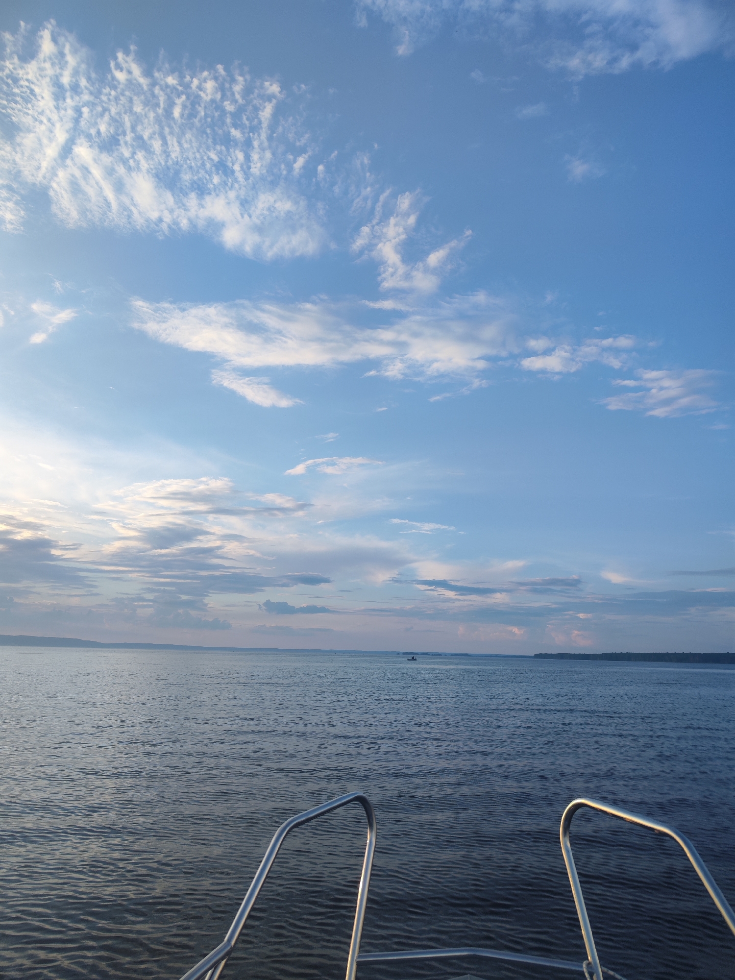 The image depicts a calm body of water, likely a lake or large river, under a partly cloudy blue sky. The perspective is from the front of a boat, with two polished metal handrails visible in the foreground. The water's surface is gently rippled, reflecting the sky. In the distance, a tree-lined shore is visible under a band of clouds tinged with the soft light of either sunrise or sunset. A small boat is present on the water in the mid-ground, to the left of the frame. The overall impression is one of peaceful tranquility. No people are directly visible in the image. The location is stated to be the city of Galevo, Russia.