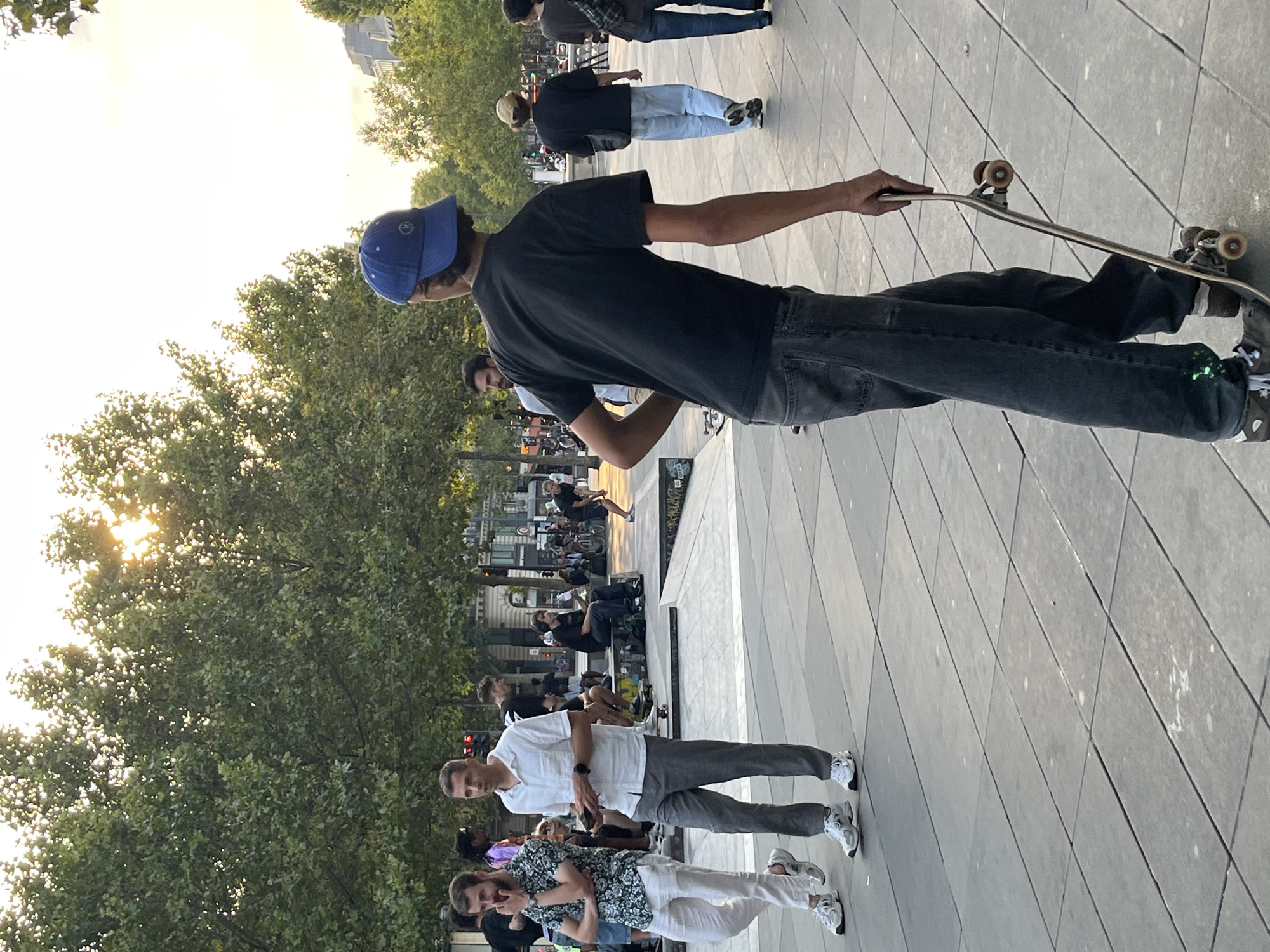 A street scene in Paris, France, features several individuals engaged in various activities. The foreground is dominated by a young man wearing a blue baseball cap and a black t-shirt, holding a skateboard. He appears to be walking away from the camera. In the middle ground, two men are walking towards the camera; one wears a printed shirt and white pants, while the other is in a white button-down shirt and gray pants. Behind them, other people are visible sitting on benches and near a skate park structure. The background is lined with trees, with the sun setting or rising, casting a warm glow and dappling sunlight through the leaves. Buildings are visible in the distance. The ground is paved with large, square tiles. The atmosphere suggests a casual outdoor gathering, possibly in a public park or urban plaza.