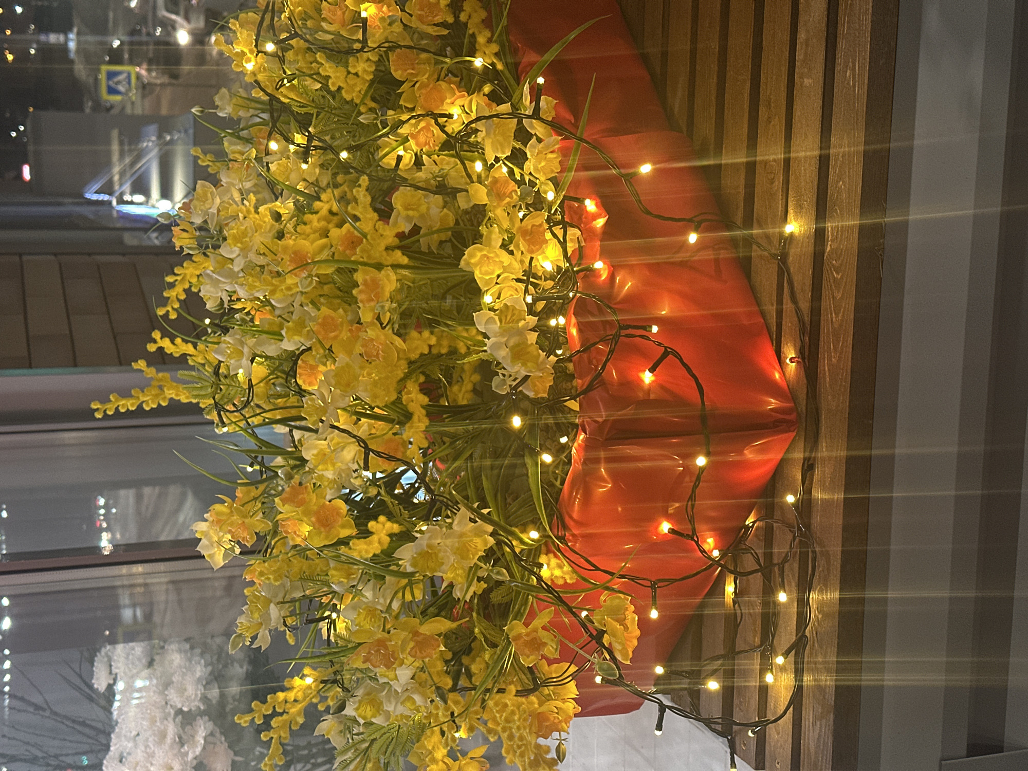 The scene depicts an indoor decorative arrangement positioned on a dark brown, horizontally slatted wooden surface, in front of a window overlooking a city street at night. A rectangular red planter, containing a dense display of artificial yellow and white flowers, resembling daffodils and mimosa, is centrally placed. A string of warm white LED lights is draped over the floral arrangement and the red planter, providing illumination and extending onto the wooden surface. Through the window, the outdoor environment is dimly visible, characterized by blurred distant lights from urban infrastructure and vehicles. A blue square pedestrian crossing sign, featuring a white human figure, is discernible through the window glass, indicating a street-level view in an urban setting. This location is identified as being within the city of Kazan, Russia.