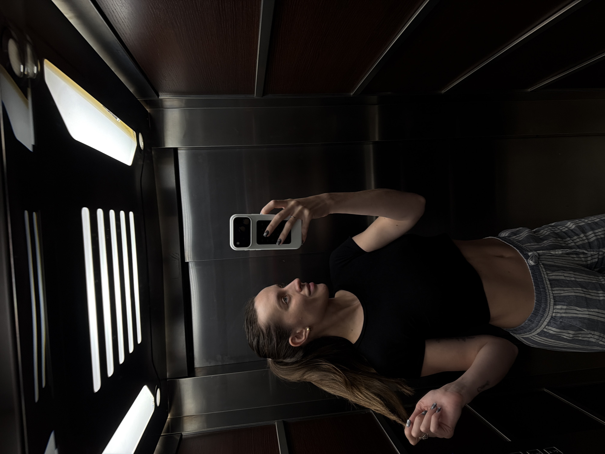 The image depicts a woman taking a mirror selfie inside an elevator, presented with a 90-degree rotation, making her appear to be lying down. The woman has long, straight brown hair tied back in a ponytail, and she wears a black short-sleeved top that exposes her midriff, paired with loose-fitting blue and white striped pants. She has dark-colored nail polish on her fingers and wears a ring on her left hand. A small gold stud earring is visible in her left ear. She holds a white smartphone with a prominent camera array, looking towards its screen with a neutral expression.
The setting is a modern elevator interior, characterized by dark, polished surfaces. The ceiling and upper wall sections feature dark brown wood panels with visible grain, separated by metallic strips. The lower walls and the surface where the woman is reflected are made of shiny, metallic material, likely stainless steel or chrome, showing reflections of the bright lights. On the left side of the frame, bright, rectangular light fixtures are prominent, providing illumination to the space. The lighting is artificial and bright, contrasting with the dark finishes of the elevator. There is no visible text in the image. The location context provided is Budva, Montenegro.