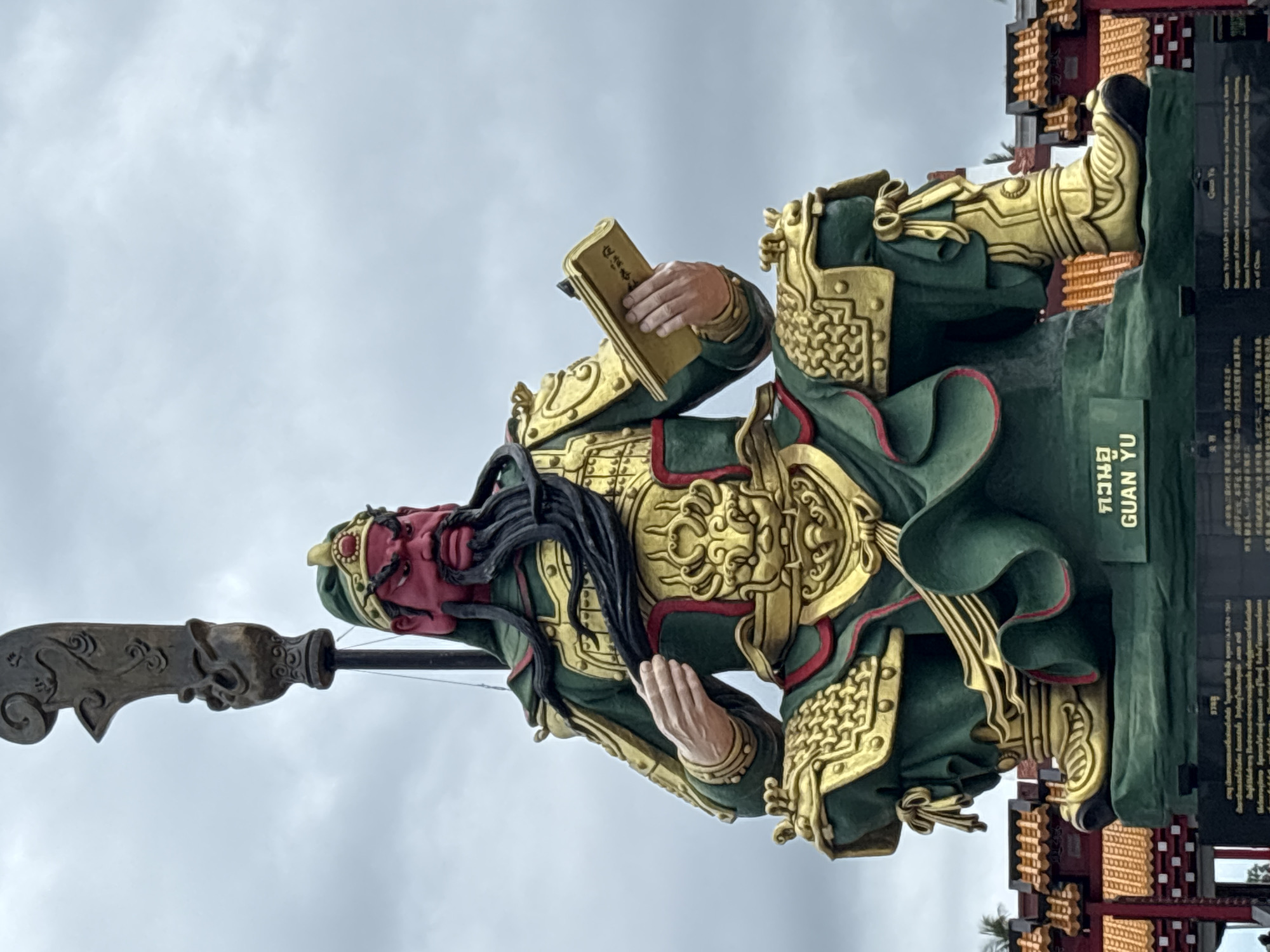The image features a statue of Guan Yu in Baan Hua Thanon, Thailand. Guan Yu is depicted with a red face, long black beard, and wearing green and gold armor. He is holding a book in one hand and appears to have a polearm weapon to his left. The statue is positioned against a cloudy sky. To the right of the statue is a plaque with text in Thai and English. The plaque mentions "GUAN YU" and includes other information.