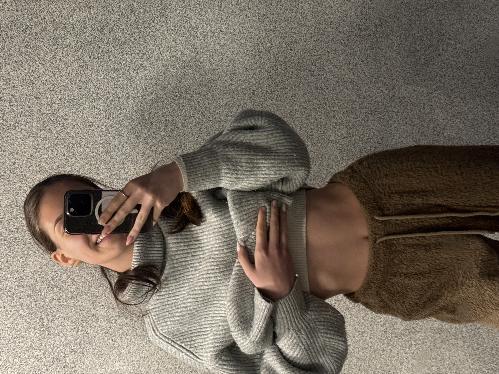 A young woman is captured in a rotated image, appearing to lie on her back on a light grey, finely textured surface, likely a carpet or floor. She holds a black smartphone with her left hand, taking a selfie. Her face, partially hidden by the phone, displays a subtle smile, and her dark hair is spread around her. She wears small hoop earrings. Her right hand rests gently on her torso.
She is dressed in a light grey, ribbed knit sweater that exposes her midriff, paired with soft, brown, possibly fleece or sherpa-like trousers featuring visible drawstrings. The smartphone's case has a white circular attachment on its back. The setting appears to be indoors, with no specific time of day or weather indicated. The woman's expression conveys a sense of happiness and relaxation. No visible text is present. This image is associated with Sofia, Bulgaria.