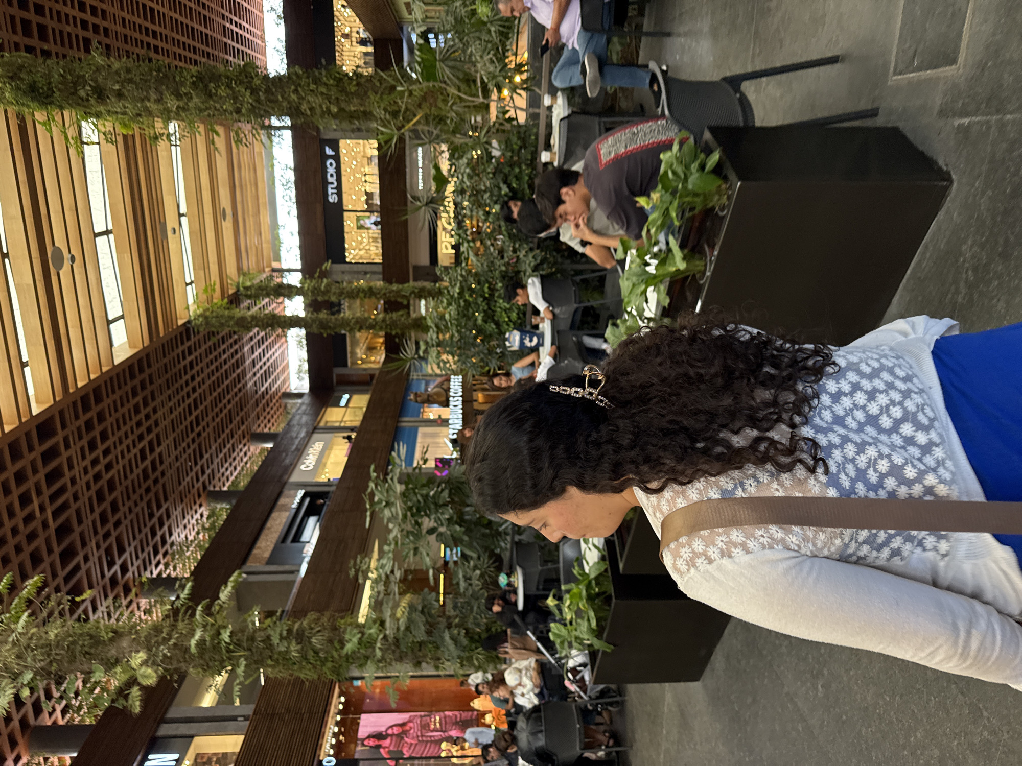 The image shows a partially-viewed woman with curly hair and a hair clip, wearing a white daisy patterned top and a blue skirt or wrap. She is sitting at an outdoor seating area in what appears to be a shopping mall or commercial center. There are other people in the background sitting at tables and walking around. Lush green plants hang from the ceiling and decorate the seating area. There are various stores in the background with recognizable branding such as "STARBUCKS COFFEE" and "STUDIO F". The architectural design of the building suggests the location is in Puebla, Mexico, based on similar known architectural styles in that city. The overall setting indicates a casual, public space where people are relaxing, shopping, or socializing.