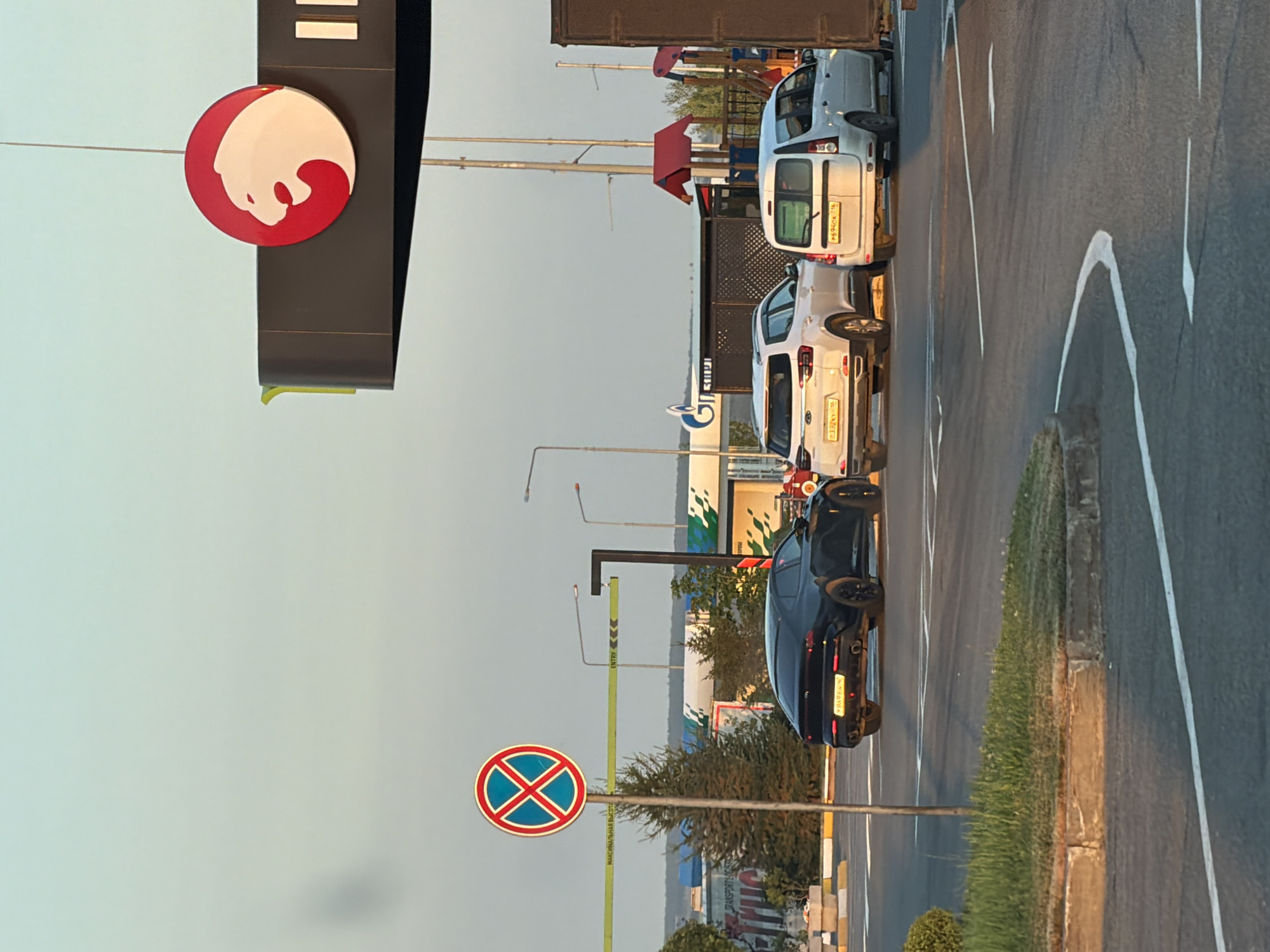 The image depicts an outdoor scene with several cars and infrastructure elements under a clear sky. In the foreground, a black sedan is parked. To its right, a white SUV and a white van are also present. A traffic sign with a red circle and a diagonal red cross is visible on the left. Above and to the right, a prominent sign with a red circular logo featuring a white lion's head is displayed. In the background, there appears to be a gas station with a Gazprom logo, and further beyond, trees and buildings are discernible. The lighting suggests it is either early morning or late afternoon.