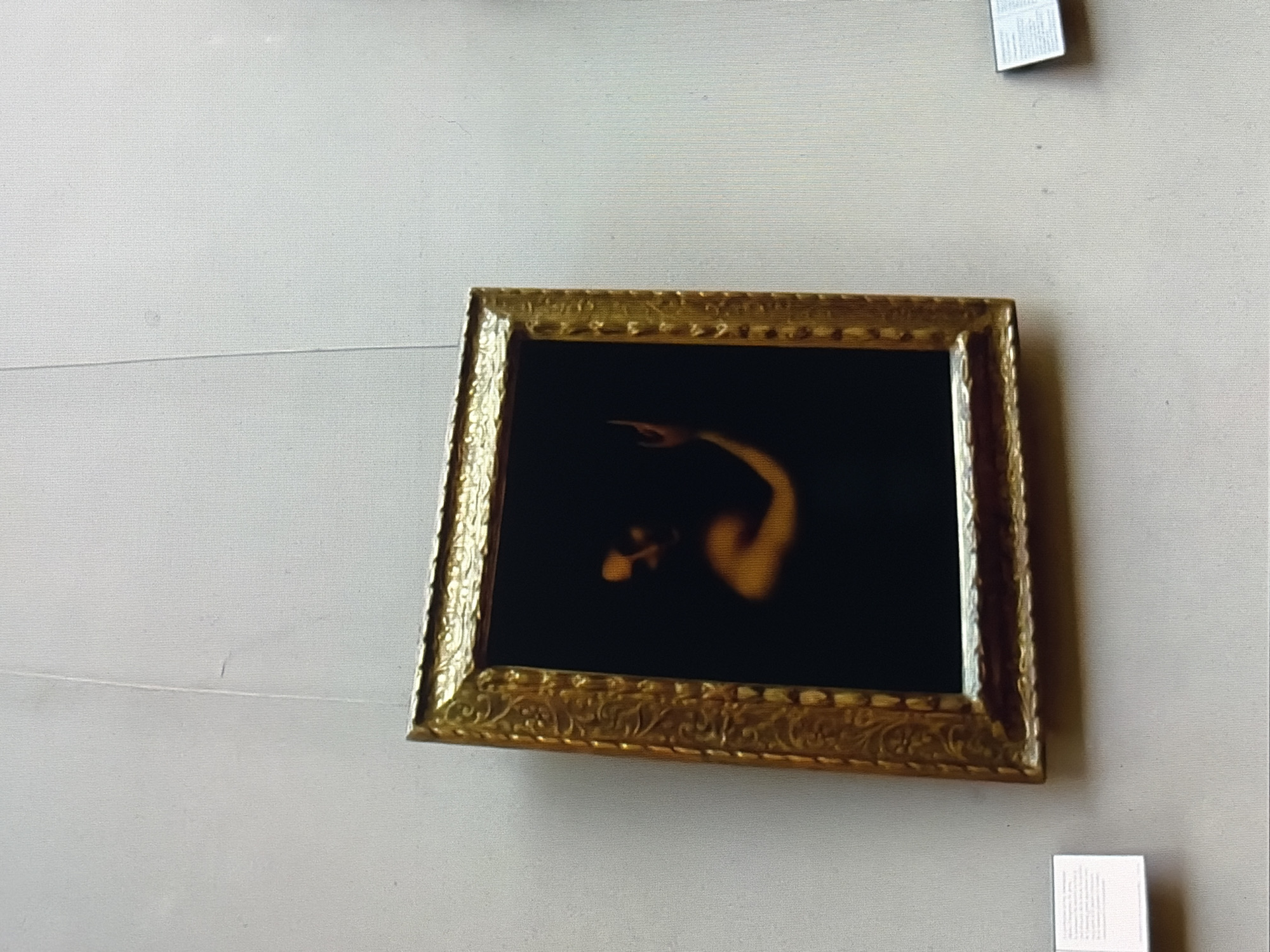 A framed painting hangs on a light-colored wall. The painting features a dark background with a figure whose skin is illuminated. The figure appears to be partially visible, with one arm raised and bent. The painting is enclosed in an ornate, gold-colored frame. To the left and right of the painting, there are small informational plaques. The overall scene suggests an art gallery or museum setting. The image is taken from a slightly angled perspective, with a subtle lean to the right.