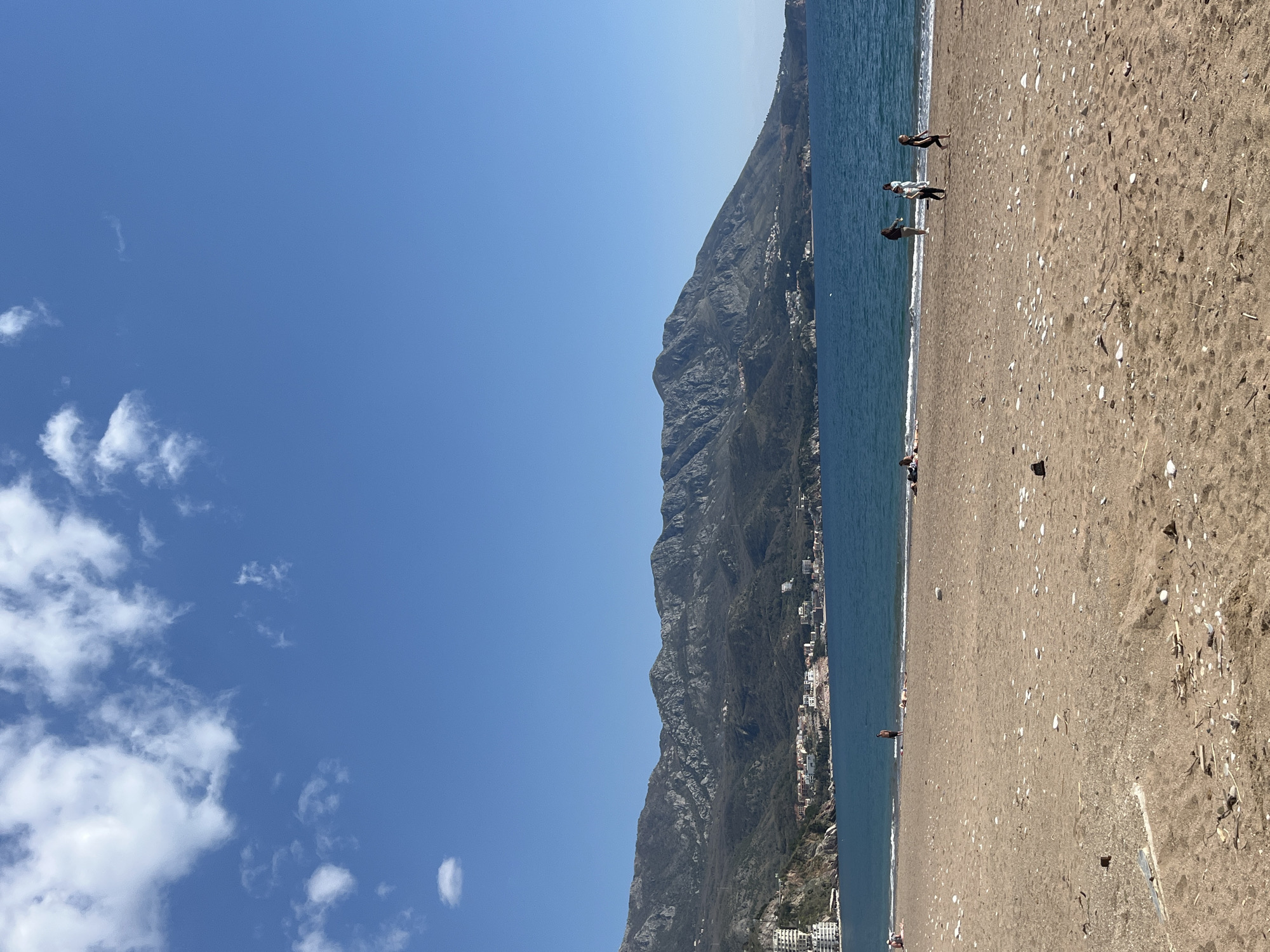 The image depicts a beach scene in Boreti, Montenegro. A steep, rocky mountain rises abruptly alongside the beach. A city is visible at the base of the mountain, along the coast. The beach has tan-colored sand with numerous shells and debris scattered across it. The water is a deep blue color. There are several people on the beach, some standing near the water's edge, and others scattered further back on the sand. The sky is blue with scattered white clouds in the left portion of the frame. The image is oriented vertically.