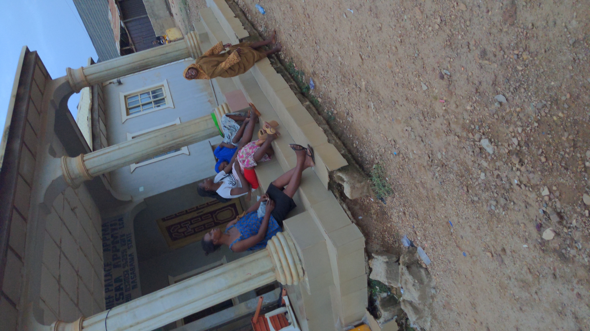 Five individuals are reclining on the steps of a building. The building is identified by signage as "Chief Palace Appymiya Saa Appymiya Iswara Appymiya LGA, Nasarawa State." The building features architectural columns and a window. The surrounding environment is a dirt ground.
