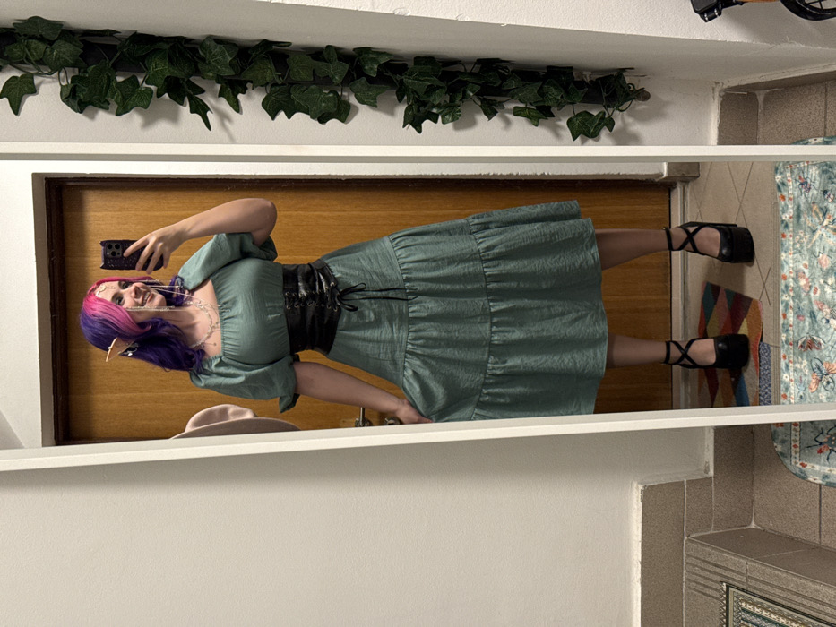 A person with purple and pink hair and elf ears poses in front of a mirror. They wear a green dress cinched with a black corset, black shoes with straps, and a pearl necklace. A hat sits on the ledge. Green ivy drapes across the top of the mirror. Reflected behind them is a wood-paneled door, a patterned rug, and a glimpse of the room's corner. They are holding a cell phone, taking their photo.