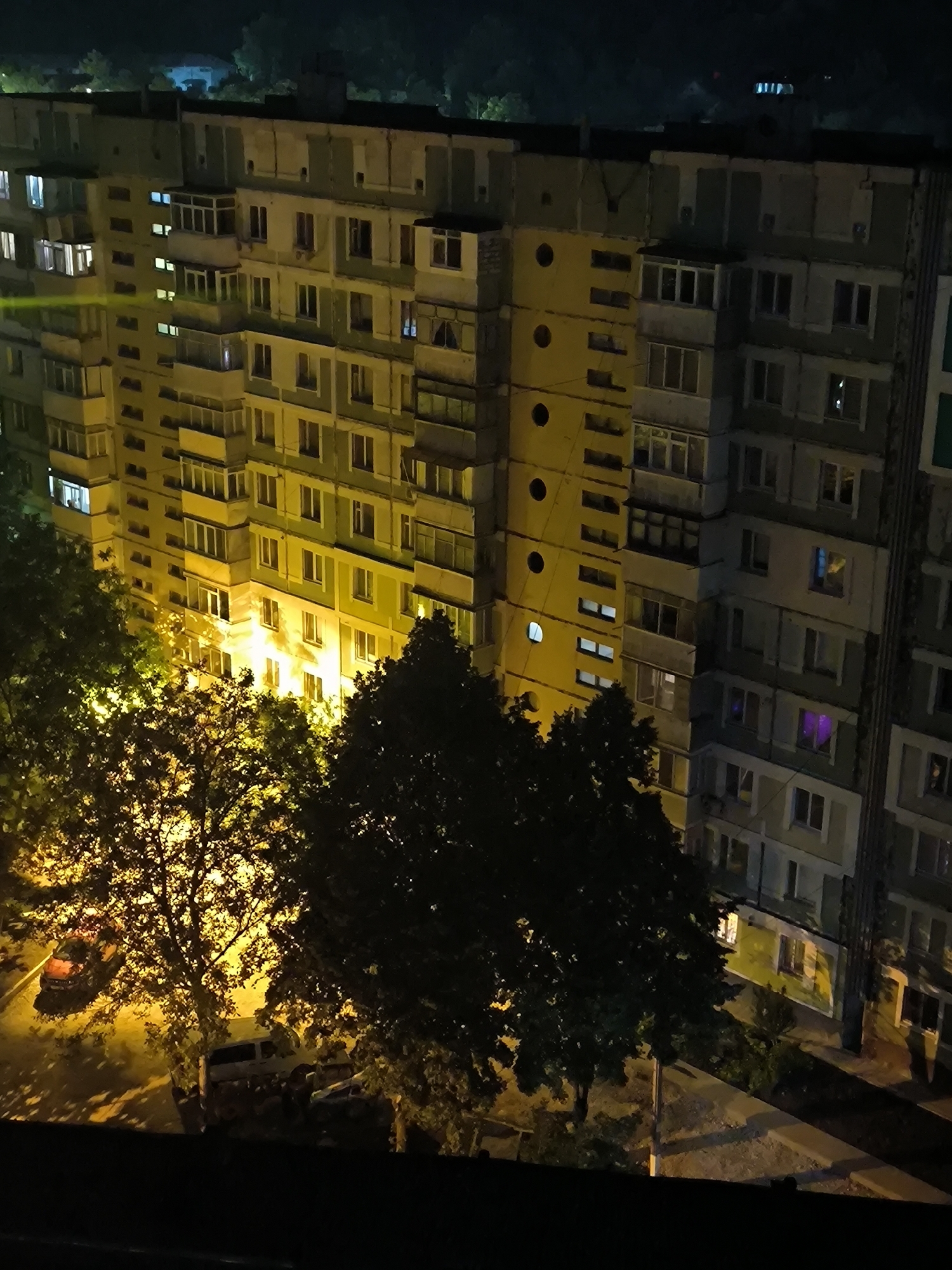 This image depicts a multi-story apartment building at night in Chișinău, Moldova. The building, likely a Soviet-era block apartment, is illuminated by yellow and white lights from its windows. Several balconies and protruding window structures are visible. In the foreground, trees obscure parts of the building and the ground below. The ground level shows parked cars, one appearing to be a white van and others possibly sedans. The overall scene suggests a residential urban environment after dark.