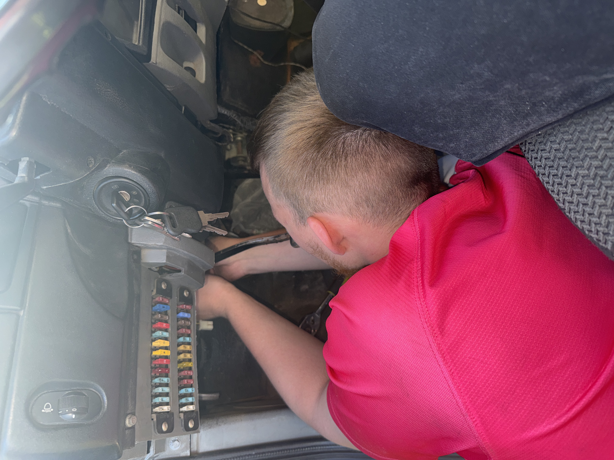 A person, appearing to be a male with short, light brown hair and a beard, is working on the fuse box of a vehicle. He is wearing a bright pink short-sleeved shirt. The fuse box is located under the dashboard, to the left of the steering column. Several fuses of various colors are visible in the fuse box. The person's head is lowered as they reach into the fuse box, holding what appears to be a tool. The steering wheel and ignition with keys are visible to the right of the fuse box. The interior of the vehicle appears to be dimly lit. There is no indication of the specific location within the city of Krasnokamsk, Russia, based on the visual information provided.