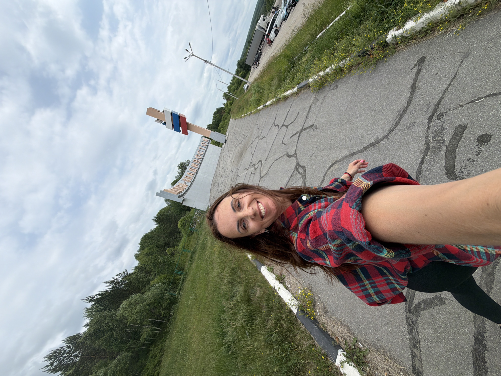 A woman in a red and black plaid shirt is taking a selfie on a cracked asphalt road. She is smiling at the camera. In the background, there is a large white sign with text in Russian that appears to say "NIKAOLAEVKA-VLADIVOSTOK". A large wooden post with three colored flags on top, red, white, and blue, is next to the sign. To the right of the road, there is a parking lot with several cars and trucks, and beyond that, a forest. The sky is cloudy and overcast.
