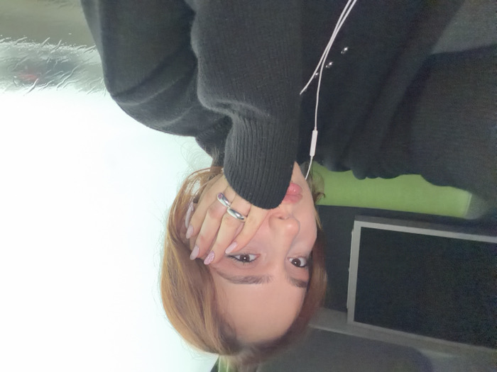 An individual is depicted in a close-up portrait inside what appears to be a vehicle in Moineville, France. The subject, a female with light brown hair, wears a dark, possibly black, long-sleeved sweater. Her left hand, adorned with two silver-colored rings on the fourth finger and light-colored manicured nails, rests against her left cheek and temple. White wired earbuds are visible, one earpiece hanging near her chest. To the subject's left, a dark rectangular screen is mounted above a bright green headrest or cushion within the vehicle's interior. To the subject's right, a window exhibits rain streaks, partially obscuring a blurred, light-toned exterior with a faint, dark-colored object, possibly a vehicle displaying red and black tones. The external conditions suggest precipitation and an overcast or foggy environment. The subject maintains direct eye contact with the camera.