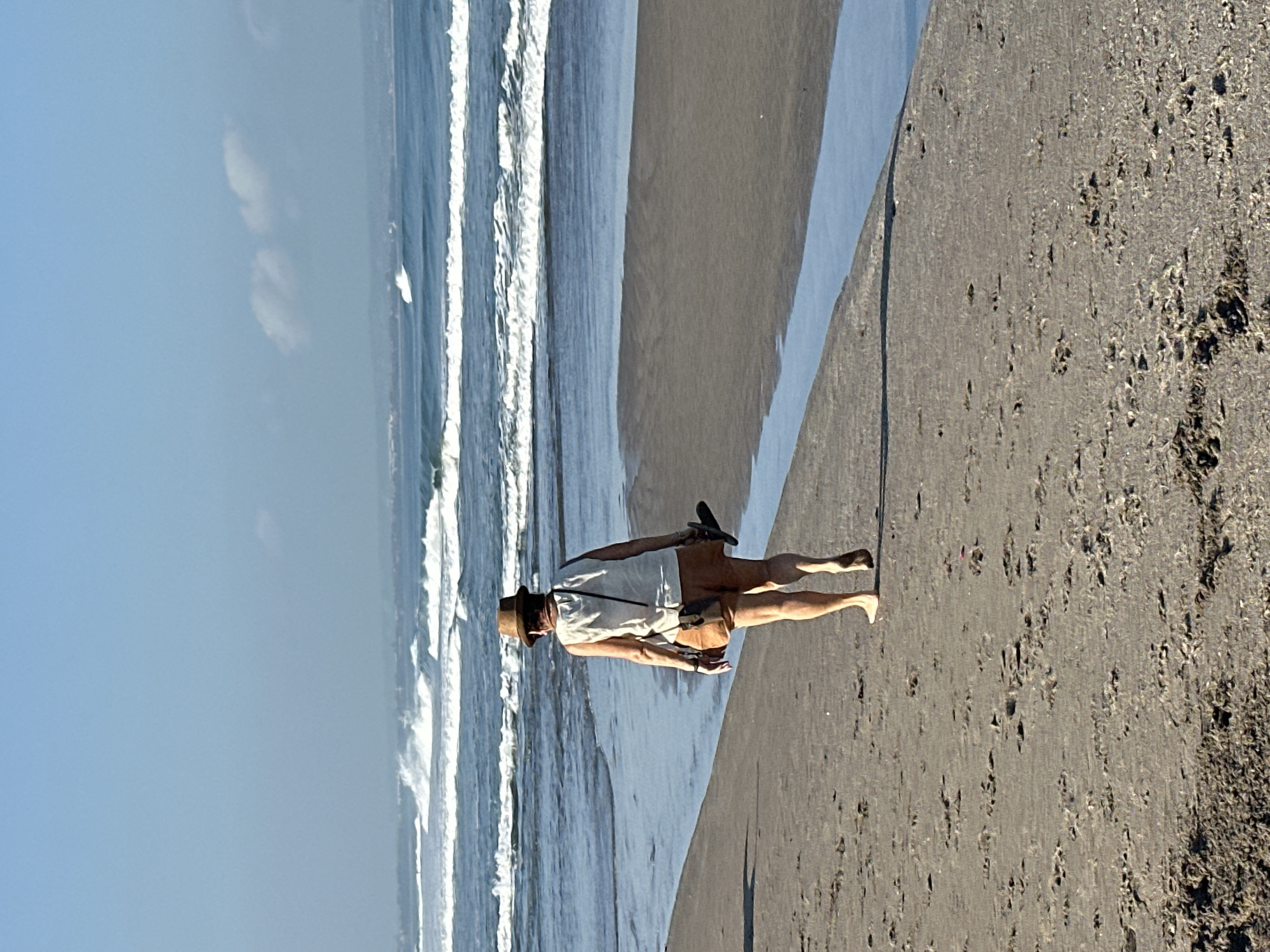 A full-shot, rear-facing, eye-level, outdoor, natural light photograph depicts a lone individual walking on a sandy beach in North Kuta, Indonesia. The person is wearing a light-colored tank top, brown shorts, and a brown fedora hat. They are carrying a pair of dark sandals in their right hand. The individual is walking towards the ocean, which features several rows of white-capped waves rolling towards the shore under a clear, blue sky with a few scattered clouds. The beach is wet and sandy, with a small stream of water running along the edge of the dry sand. The overall scene conveys a sense of tranquility and relaxation.
