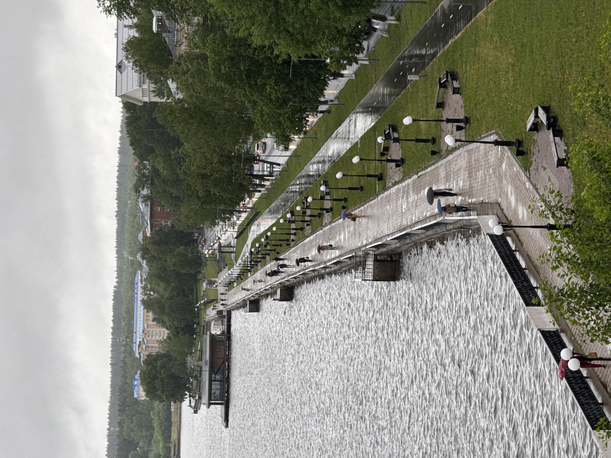 The image depicts a waterfront promenade in Votkinsk, Russia, on a rainy day. The promenade, constructed of light-colored paving stones, runs parallel to a body of water, likely a lake or reservoir. A row of evenly spaced, spherical white lampposts lines the water side of the promenade. Along the opposite side, a strip of green lawn separates the promenade from a street with vehicle traffic and a row of trees. People are walking along the promenade, some carrying umbrellas, suggesting the presence of rain. The sky is overcast, reinforcing the rainy weather conditions. Buildings and wooded hills are visible in the distance on the opposite side of the body of water. The water appears choppy due to the weather. Several metal staircases provide access to the water from the promenade. Benches are placed on the lawn beside the walkway, adjacent to the light posts.