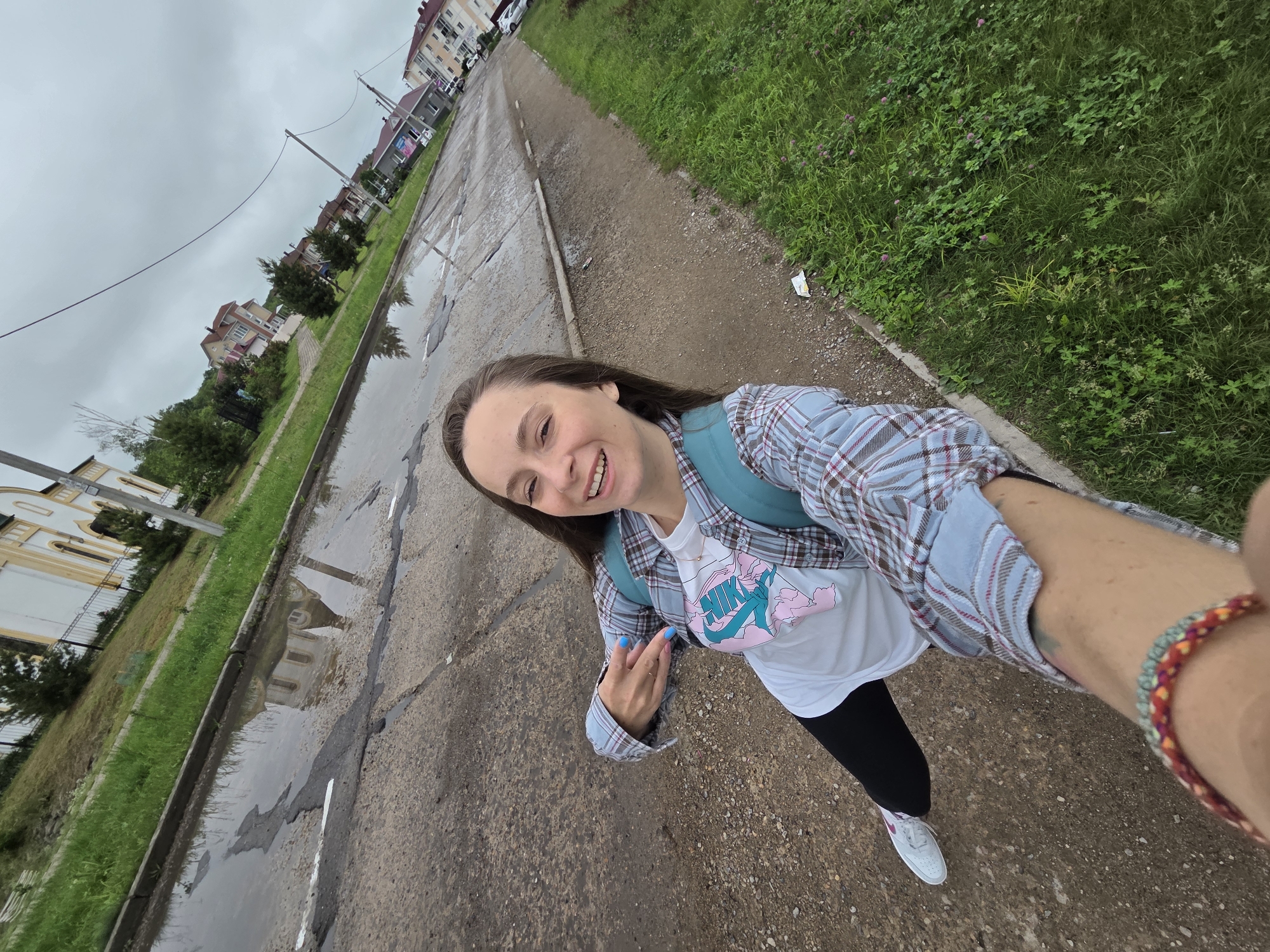 A full shot from a high angle shows a woman standing on a dirt path next to a paved road with puddles and potholes. The woman is wearing a blue and white plaid shirt, a white t-shirt with a graphic print, black leggings, and white sneakers. She has a teal backpack on. She is smiling and holding her phone in her outstretched arm, taking a selfie. The background shows buildings, trees, and a cloudy sky. The location is identified as Birobidzhan, Russia.