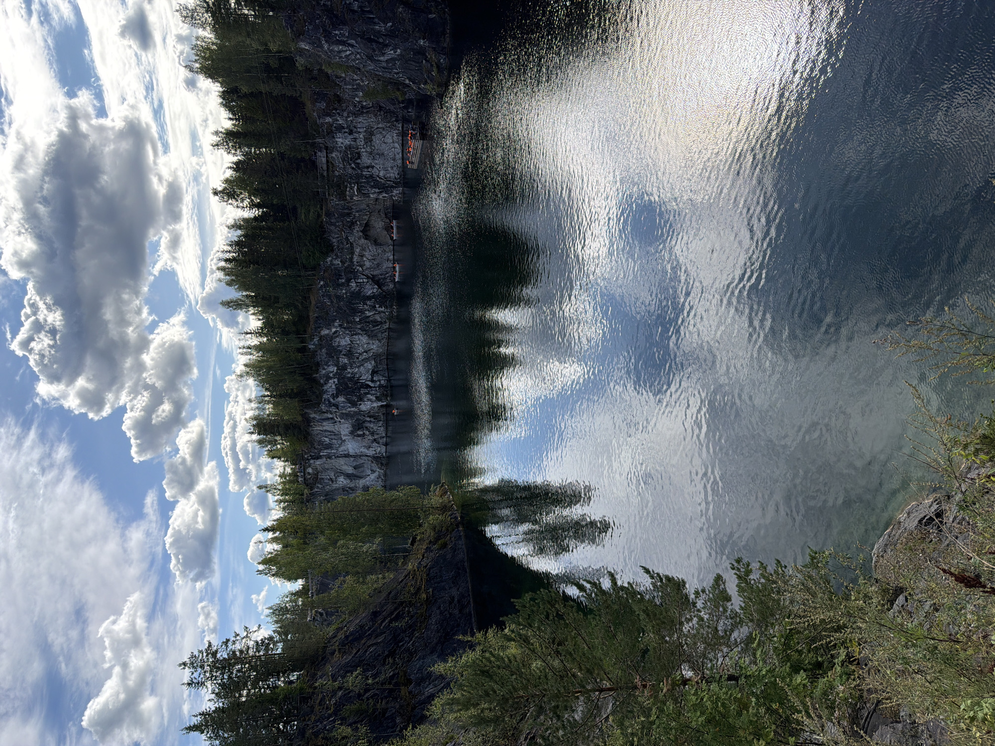 This is an outdoor, vertical, eye-level shot of a deep, dark body of water surrounded by sheer, grey rock cliffs and dense, green trees under a bright, cloudy sky. The water reflects the sky and the trees, creating a rippling, dark mirror. Several small, wooden boats with people wearing life jackets are visible on the water. The foreground shows a rocky, tree-covered embankment. The overall scene suggests a natural landscape, possibly a quarry or a canyon.