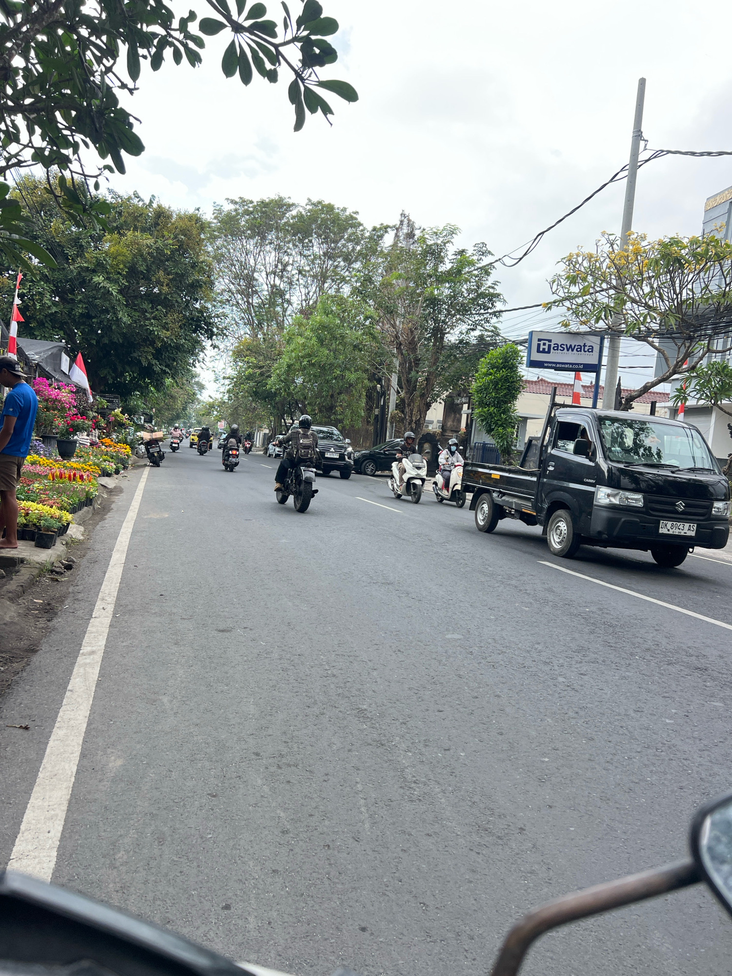 The image depicts a street scene in Denpasar, Indonesia, likely a commercial area given the presence of shops and a busy road. A black Suzuki Carry pickup truck is prominently featured in the right half of the frame, driving on the asphalt road. It has a cargo bed and a license plate reading "DK 8943 AS". Several motorcycles are also visible on the road, with riders wearing helmets. In the foreground, on the left side of the road, there is a stall selling a variety of colorful potted flowers and plants. A man wearing a blue t-shirt and brown shorts stands beside the flowers. Trees line the sides of the street, and buildings are visible in the background. A sign for "Aswata" insurance is also visible. The sky is overcast, suggesting cloudy weather. The perspective suggests the photo was taken from a moving vehicle, possibly another motorcycle.