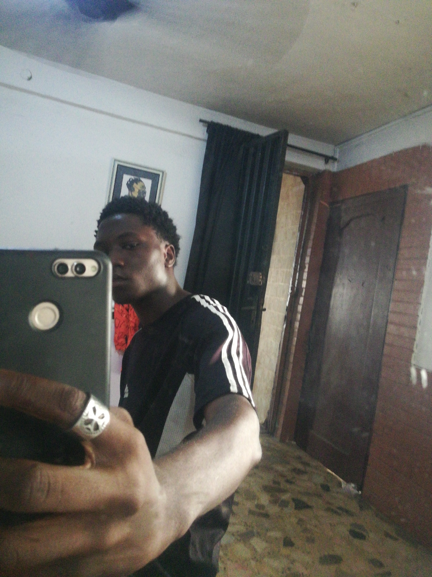 A young man is taking a selfie in what appears to be an indoor environment. He is holding a smartphone in his left hand, obscuring part of his face. He is wearing a dark shirt with white stripes on the sleeve. On his finger, there is a silver ring with a decorative design. Behind him, there is a framed picture on the wall, black curtains, and a closed wooden door with a brown frame. The floor has a mottled brown and gray pattern. The location is potentially in Lagos, Nigeria, but it is difficult to confirm without additional details.