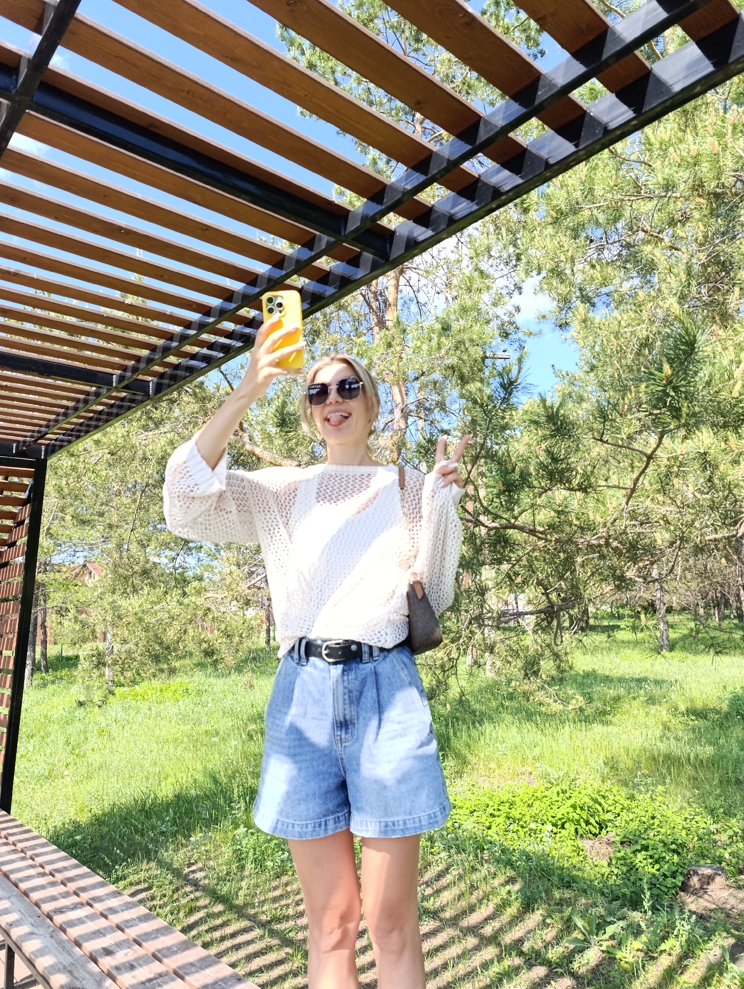 A woman stands under a pergola, taking a selfie with her phone. She is wearing sunglasses, a white sweater, and denim shorts with a black belt. She is sticking her tongue out and holding up a peace sign. The background consists of trees and green grass, suggesting a park-like environment. A wooden bench is partially visible on the left.