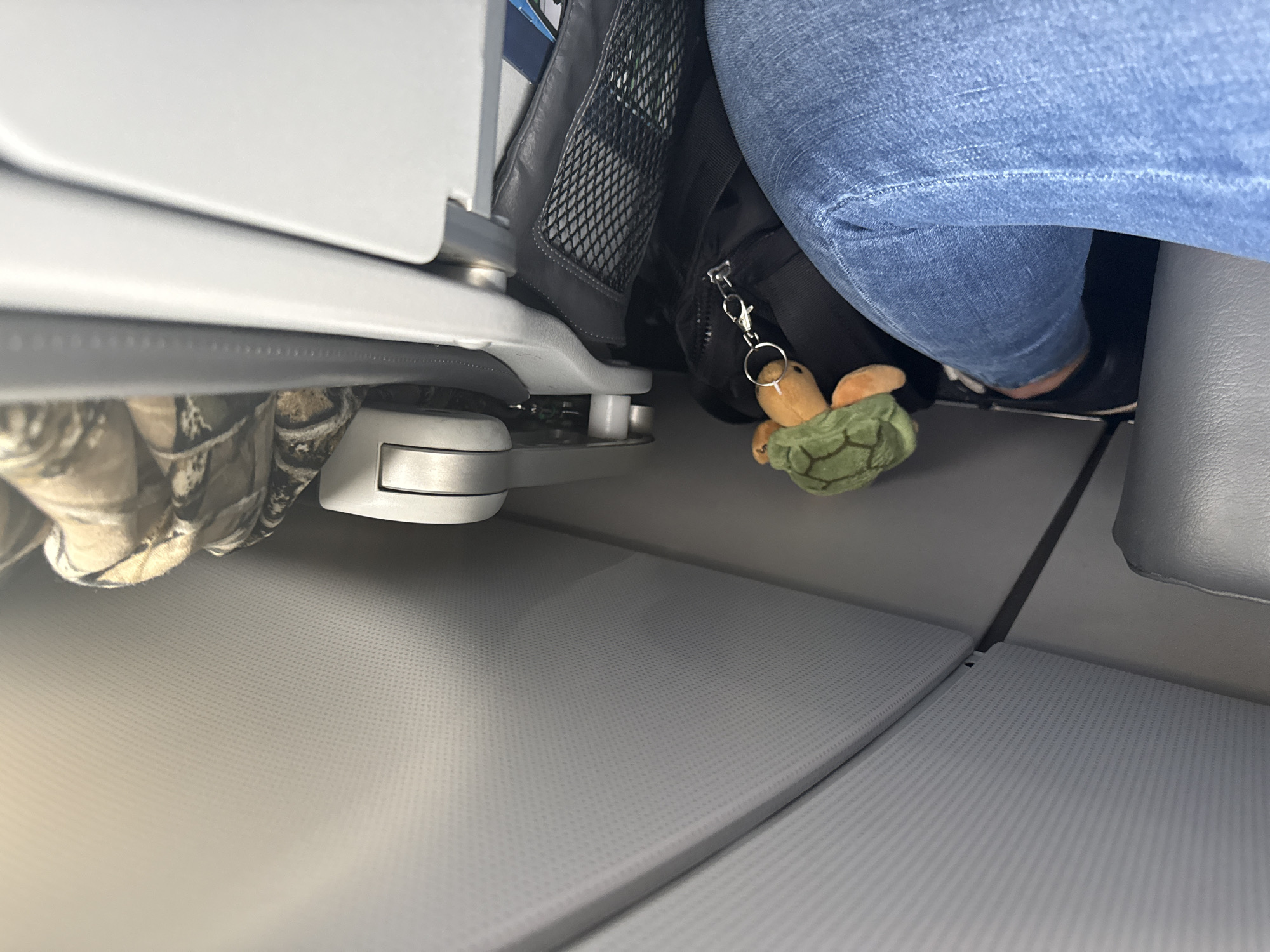 The image depicts the under-seat area of an airplane, likely during a flight or boarding process. The scene includes an airplane seat leg, a bag, and a person wearing jeans. Hanging from the bag is a plush toy turtle attached to a keychain. The flooring is a textured gray material, standard for airplane cabins. There is no information about the city of San Antonio or specific location within it.