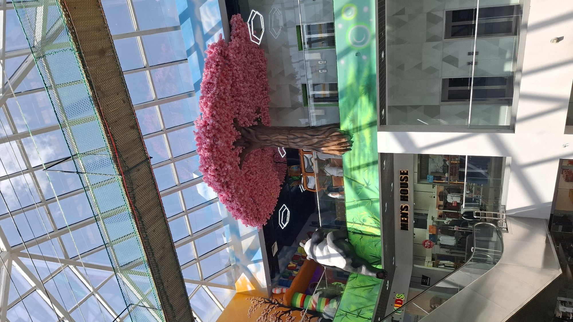 This is a full shot of the atrium of a shopping mall in Shymkent, Kazakhstan, under a glass ceiling with a visible structural grid. In the upper portion of the frame, a suspended net bridge spans the atrium, illuminated by sunlight. Below this, a large, artificial tree with vibrant pink foliage dominates the central area, appearing to grow out of a raised platform. A section of an inflatable slide, featuring colorful stripes, is visible to the left of the tree. A large, decorative panda figure is perched on the edge of the platform, looking to the left. On the same level as the panda and tree, there is a shop with the sign "MEN'S HOUSE" visible. Below this, an escalator descends, with a glass railing. Further down, another section of the mall's interior is visible, with shops and elevator doors. The overall environment suggests a brightly lit, modern indoor public space, possibly with a play area integrated into the mall's design.