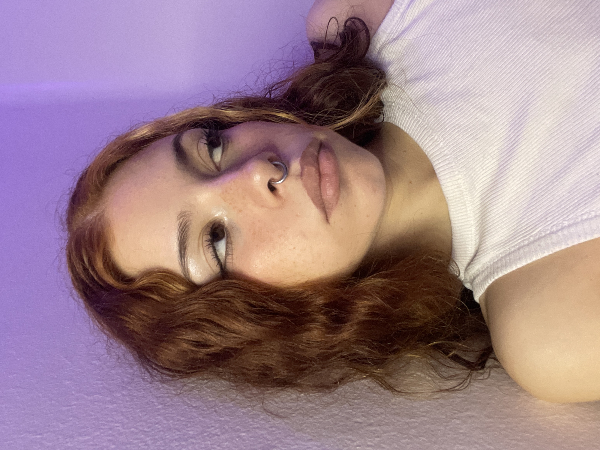 A close-up, angled view depicts a young woman with auburn hair, fair skin, and freckles. She has a piercing in her left nostril. She is wearing a white, ribbed tank top and is lying on a surface that appears to be textured and lit with a faint purple light. The location and city of Buenos Aires, Argentina, cannot be determined from the image.